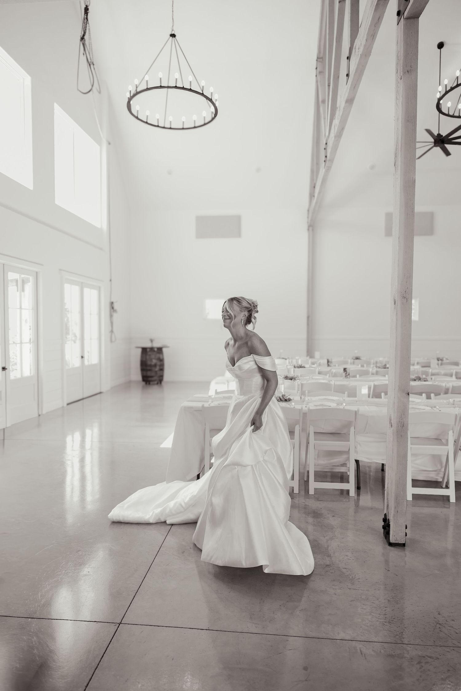 Black and white photo of a bride in an off-shoulder gown standing in an elegant white wedding venue with high ceilings.