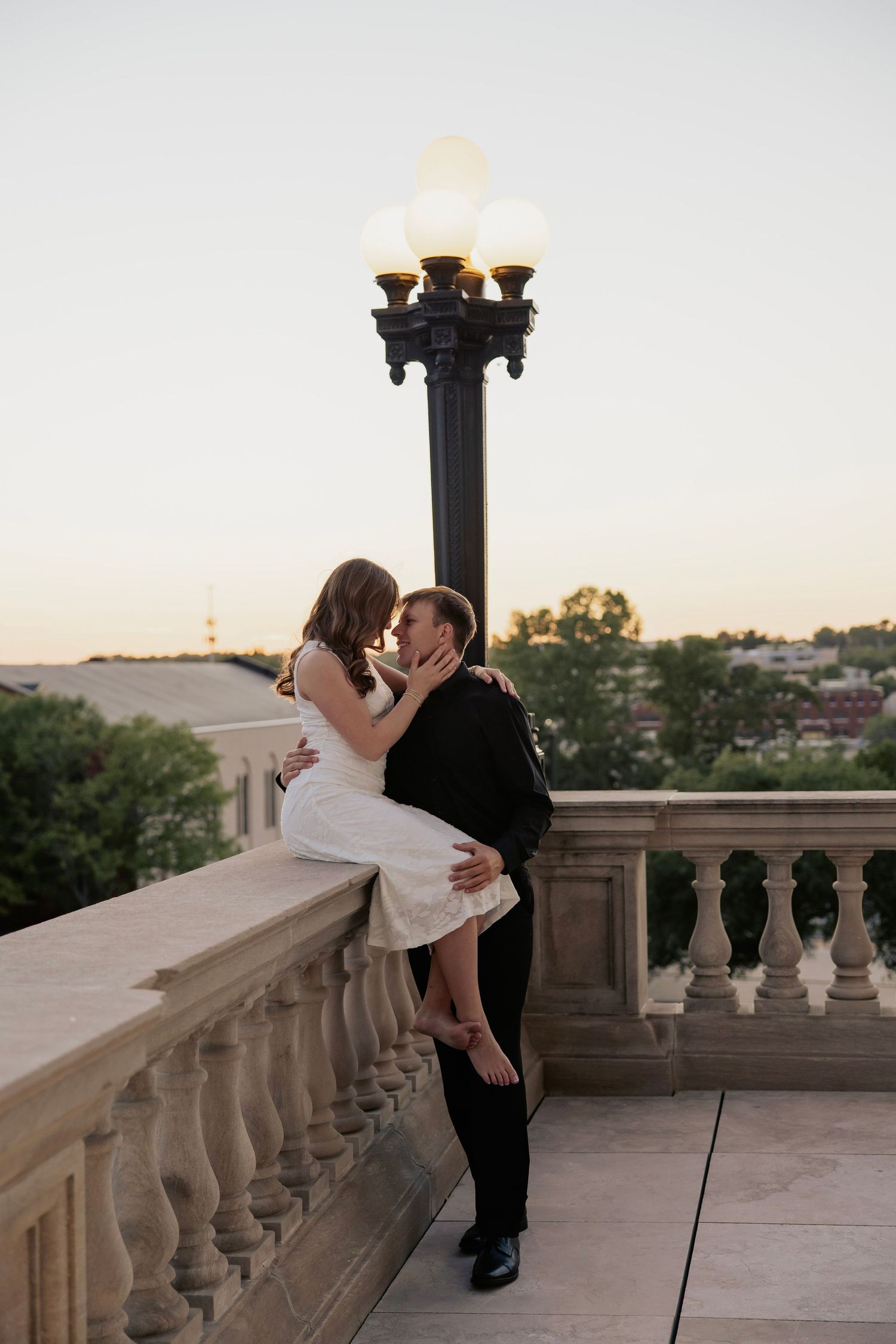 Couple sharing a romantic kiss at sunset on a stone balcony with a vintage lamp post glowing behind them.