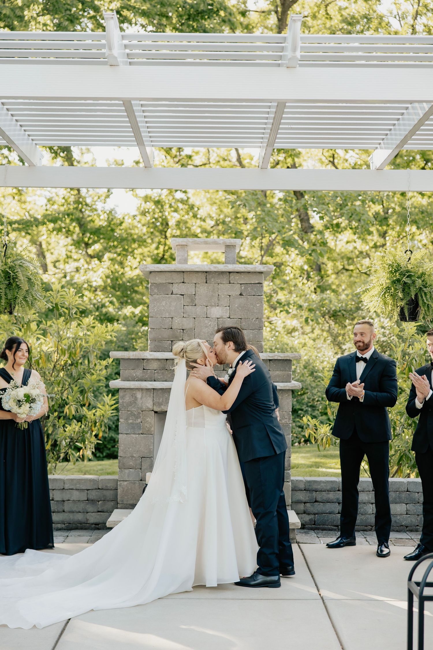 Bride and groom share first kiss under white pergola at outdoor wedding ceremony with bridesmaids and officiant nearby.