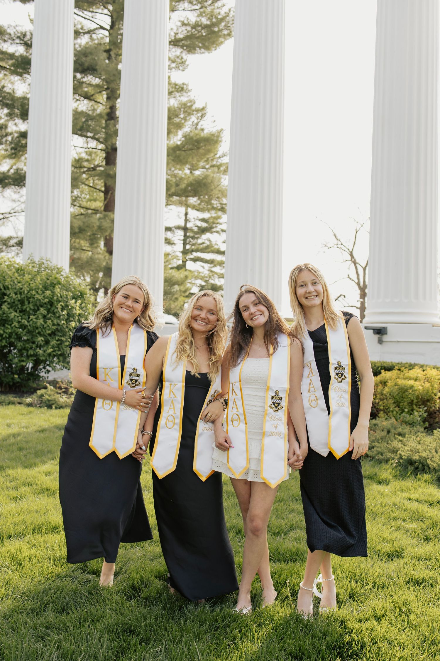 Four smiling female graduates wearing gold honor stoles posing together on green lawn.
