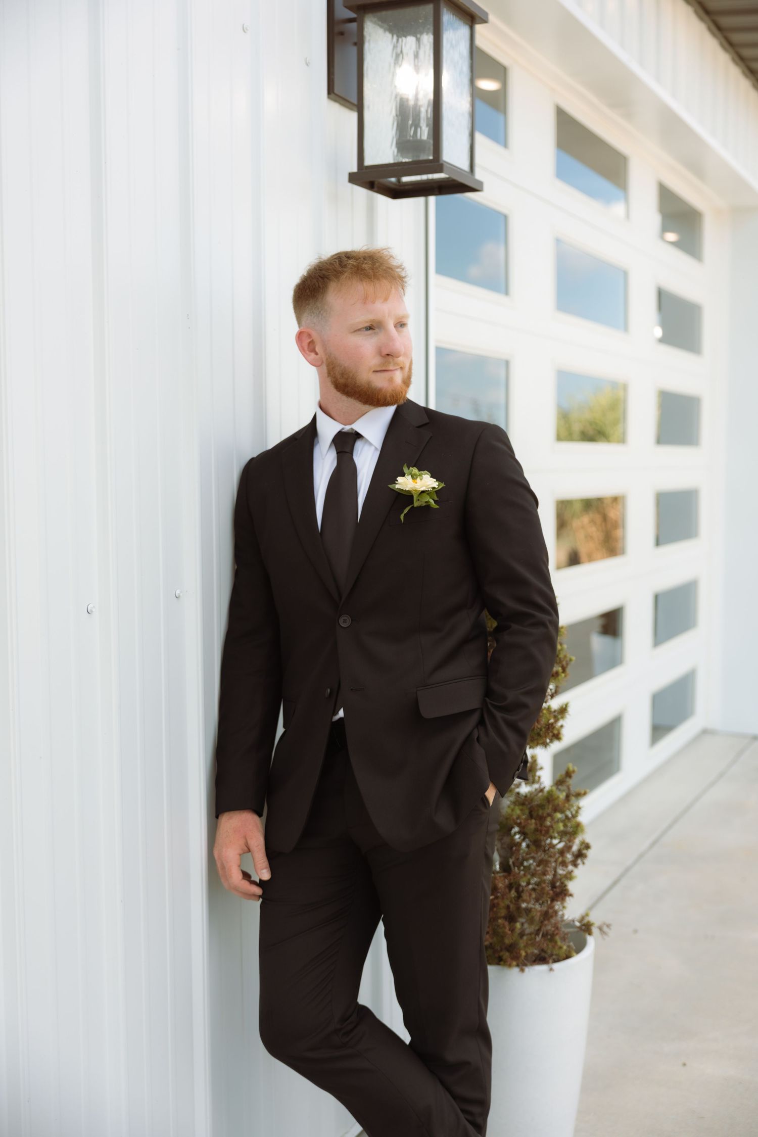 Groom in dark suit with boutonniere leans against white barn wall, looking away thoughtfully.