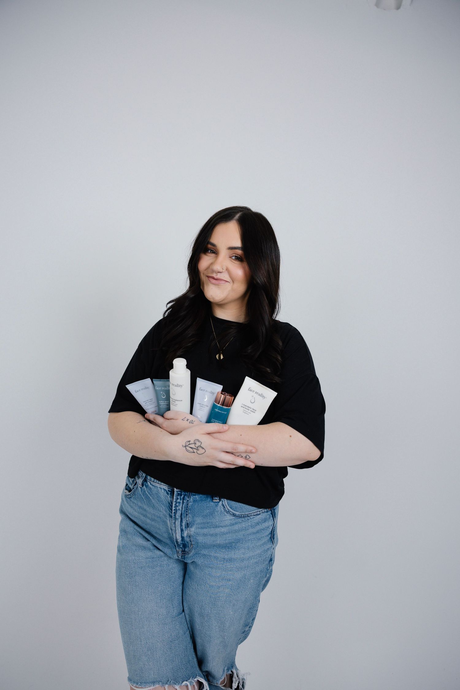 Smiling dark-haired woman in black top and jeans holding multiple skincare product tubes.