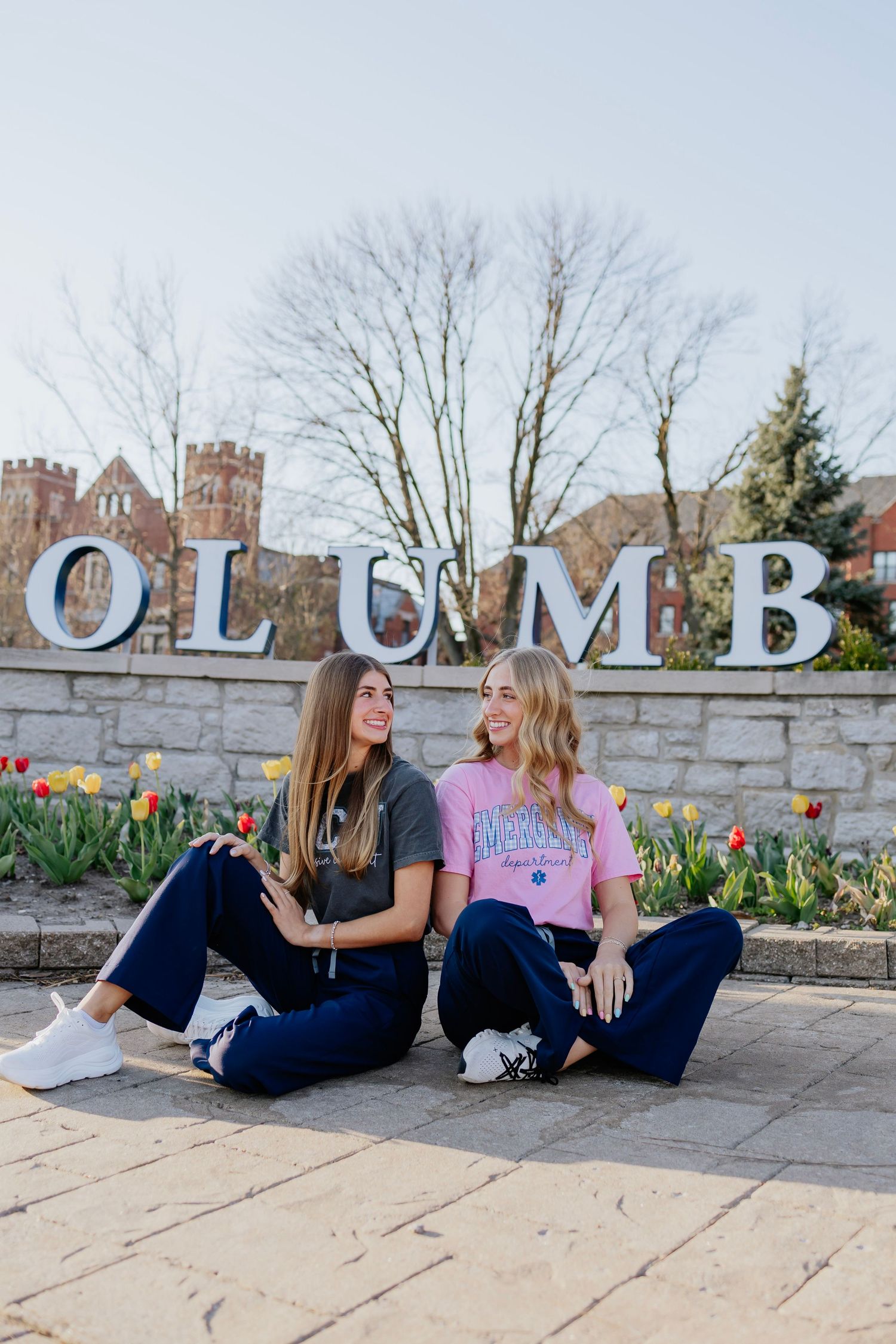 Two smiling young women sitting together in front of Columbus city sign with spring tulips.