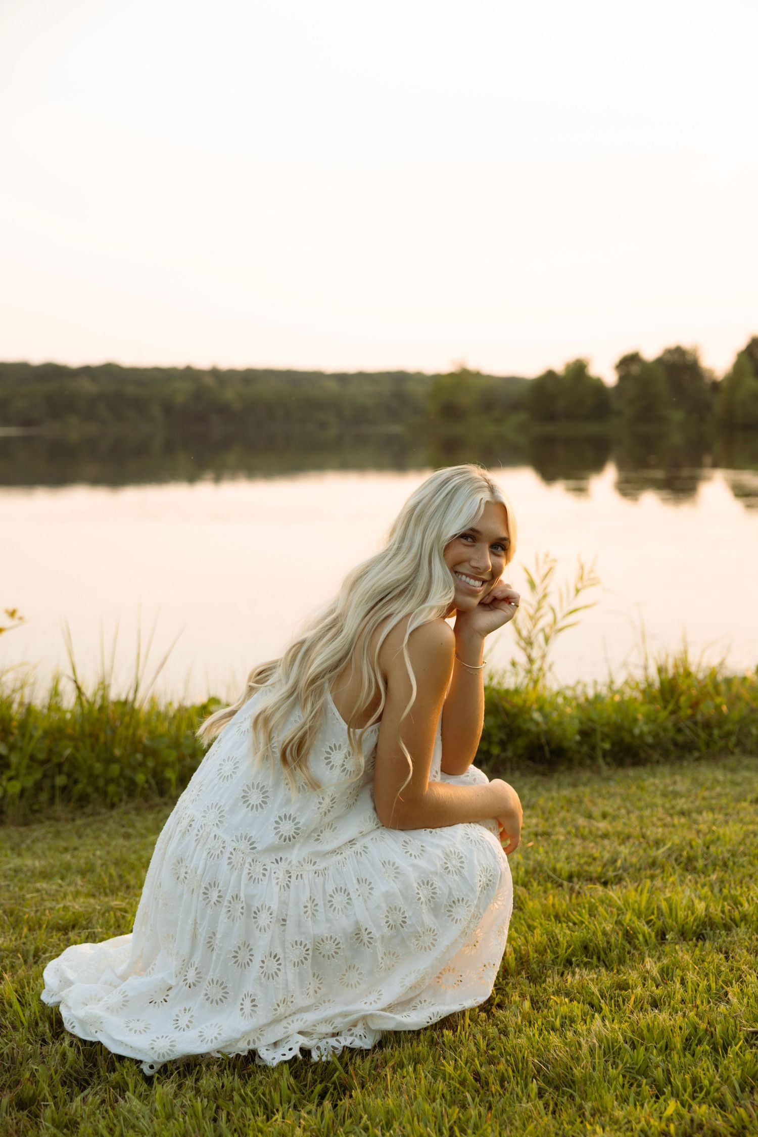 Smiling blonde woman in white lace dress crouching by a serene lake at golden hour sunset.