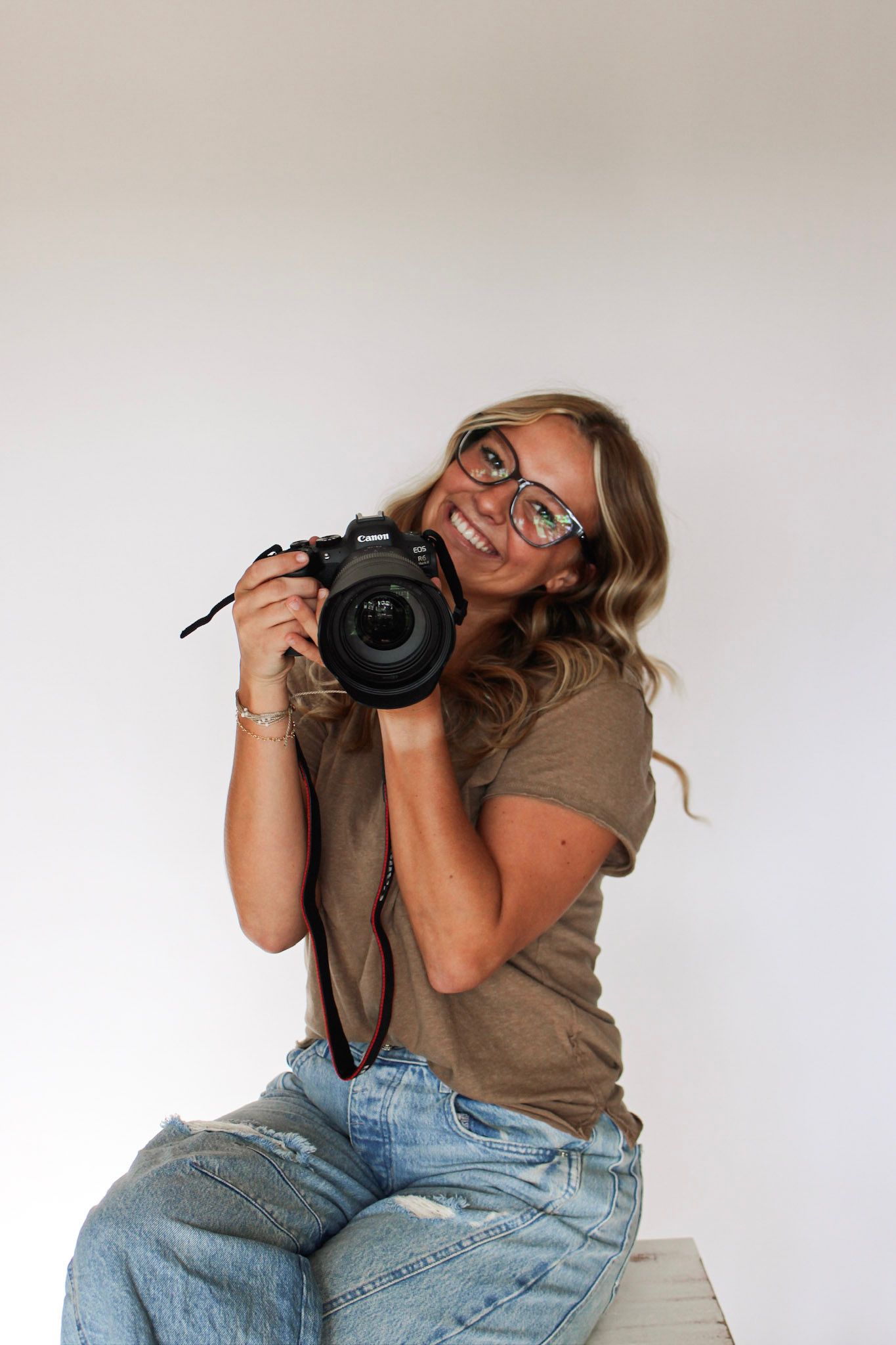 Smiling blonde woman with glasses holding Canon DSLR camera while sitting on white box.