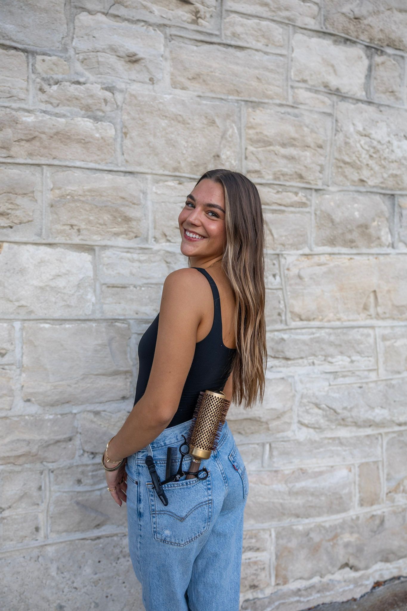 Smiling young woman in black bodysuit and Levi jeans with hairstyling tools tucked in pocket by stone wall.