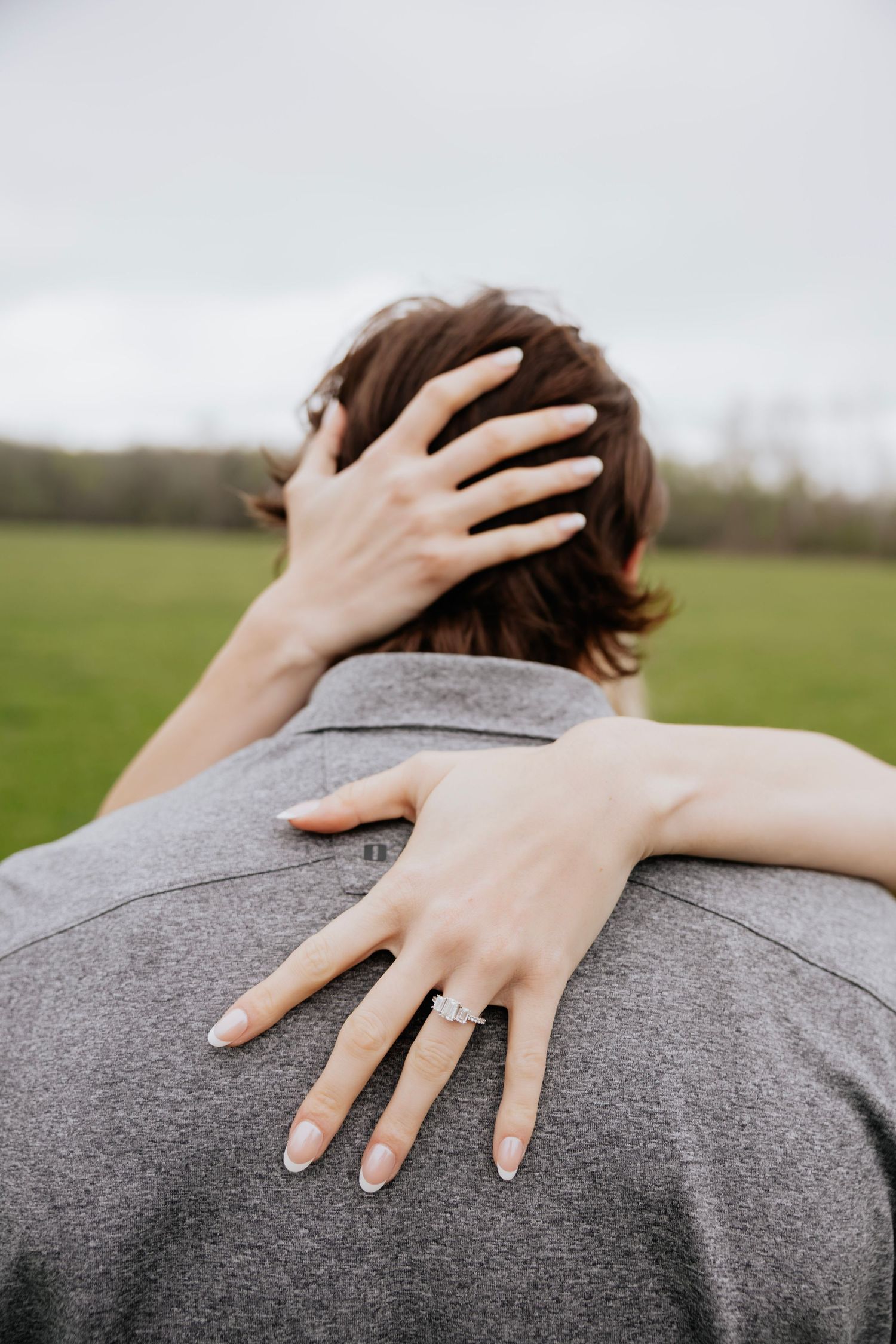 Close-up of woman's hand with diamond engagement ring resting on man's shoulder outdoors.