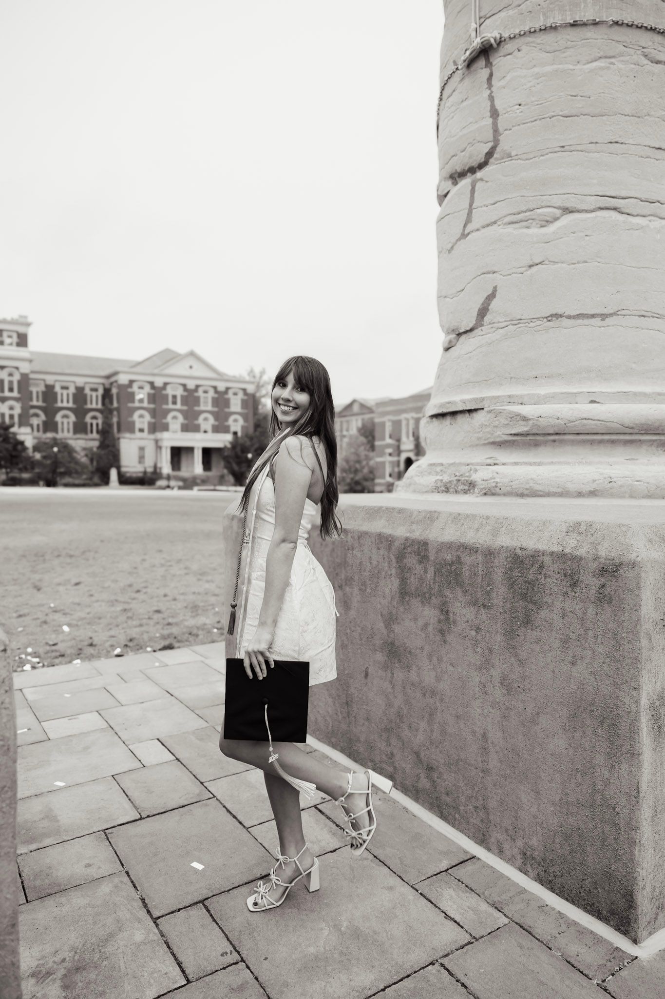 Black and white photo of smiling woman in white dress near campus column.
