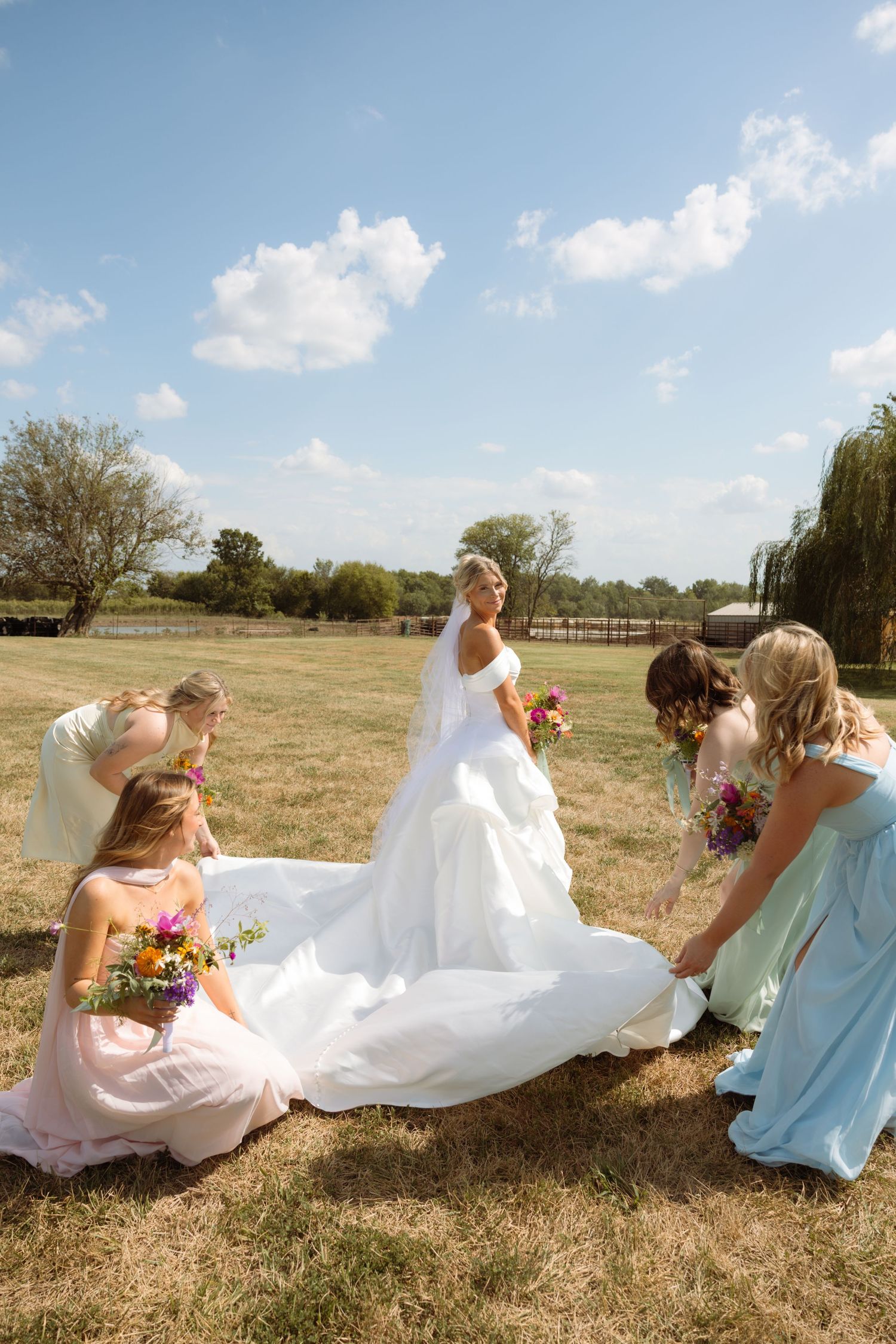 Bridesmaids arranging the train of a white wedding gown outdoors on a sunny day in a green field.
