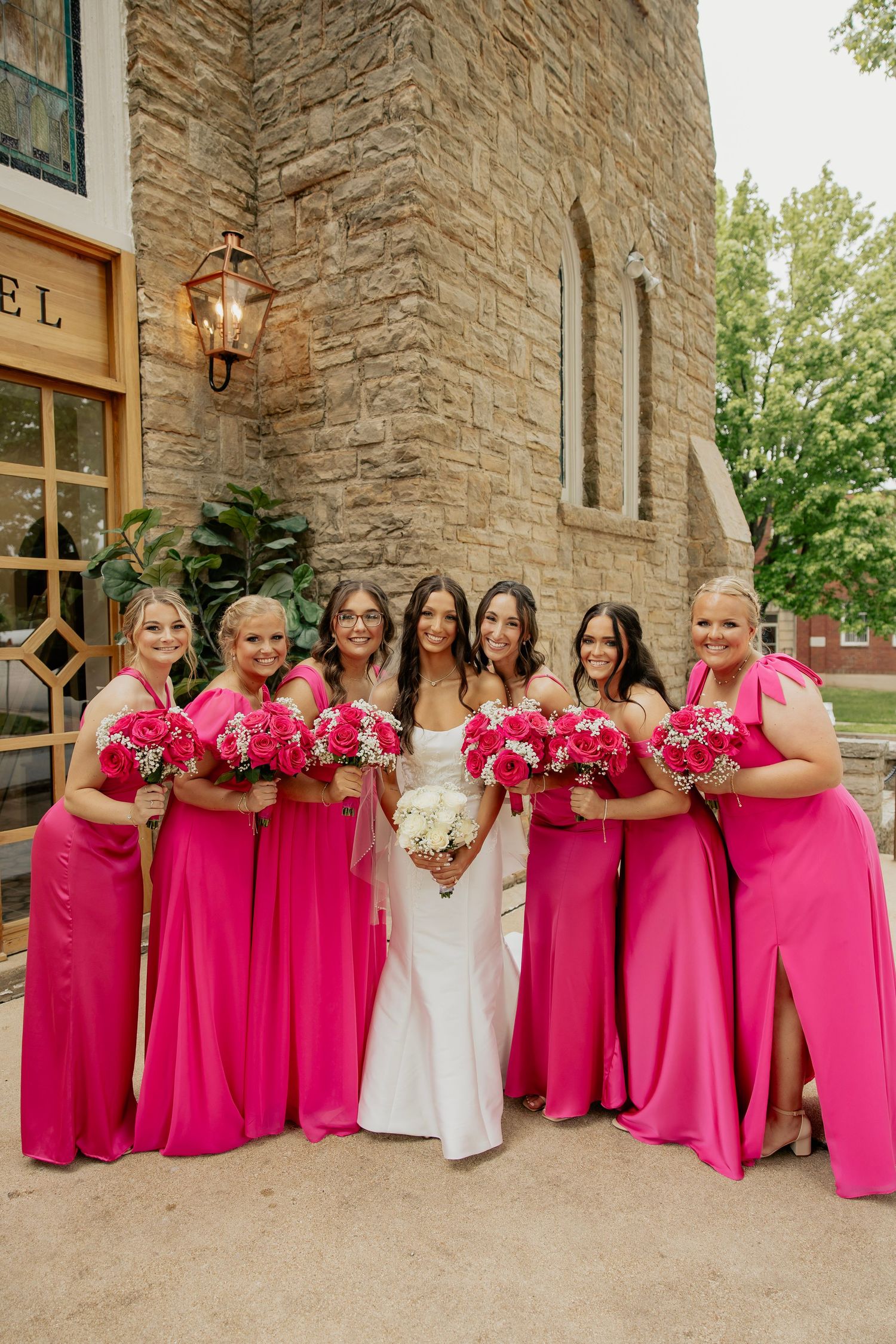 Bride in white gown with bridesmaids in hot pink dresses holding rose bouquets outside stone church.
