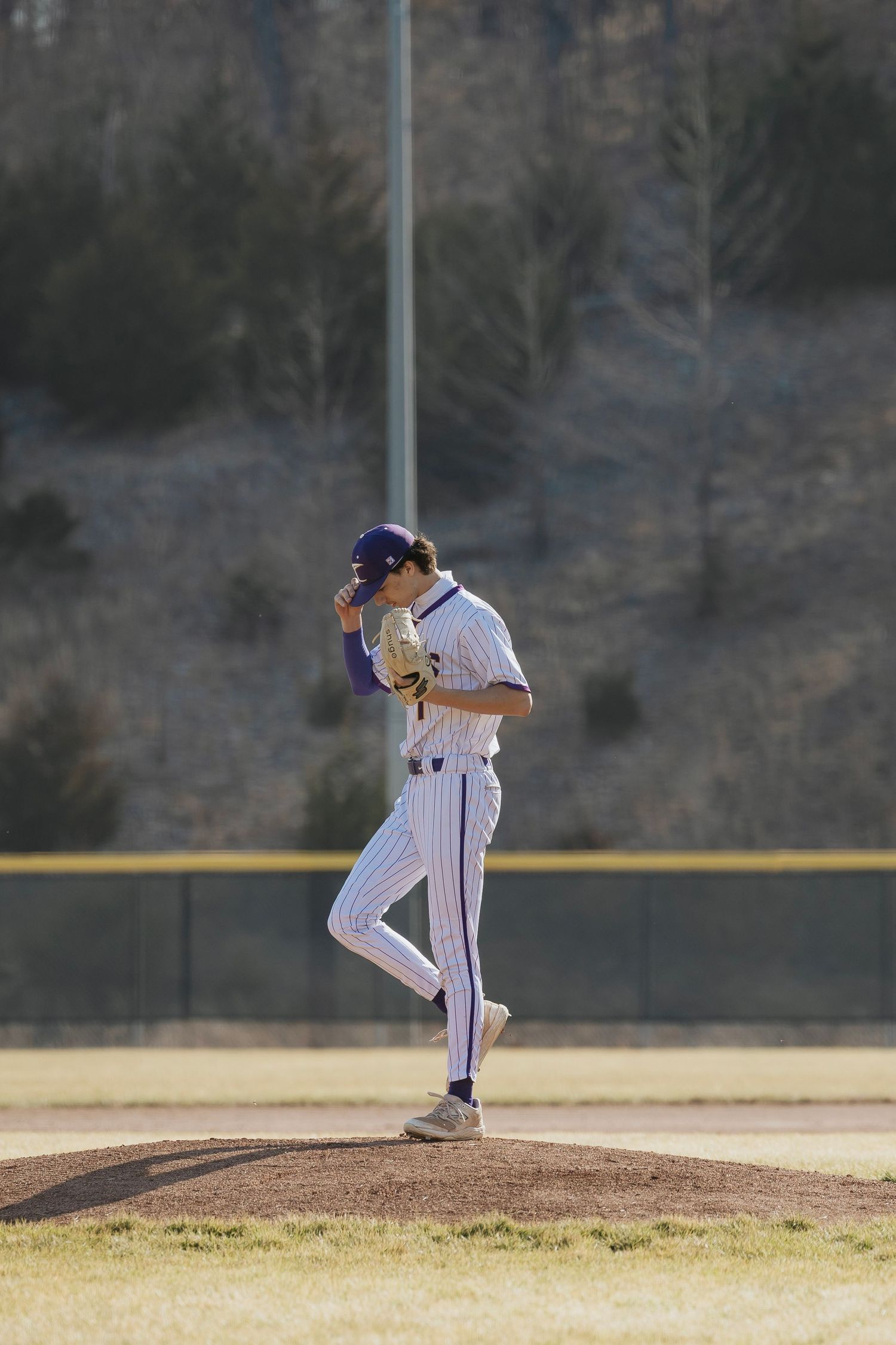 High school baseball pitcher in white pinstripe uniform winding up on the mound during a sunny outdoor game.