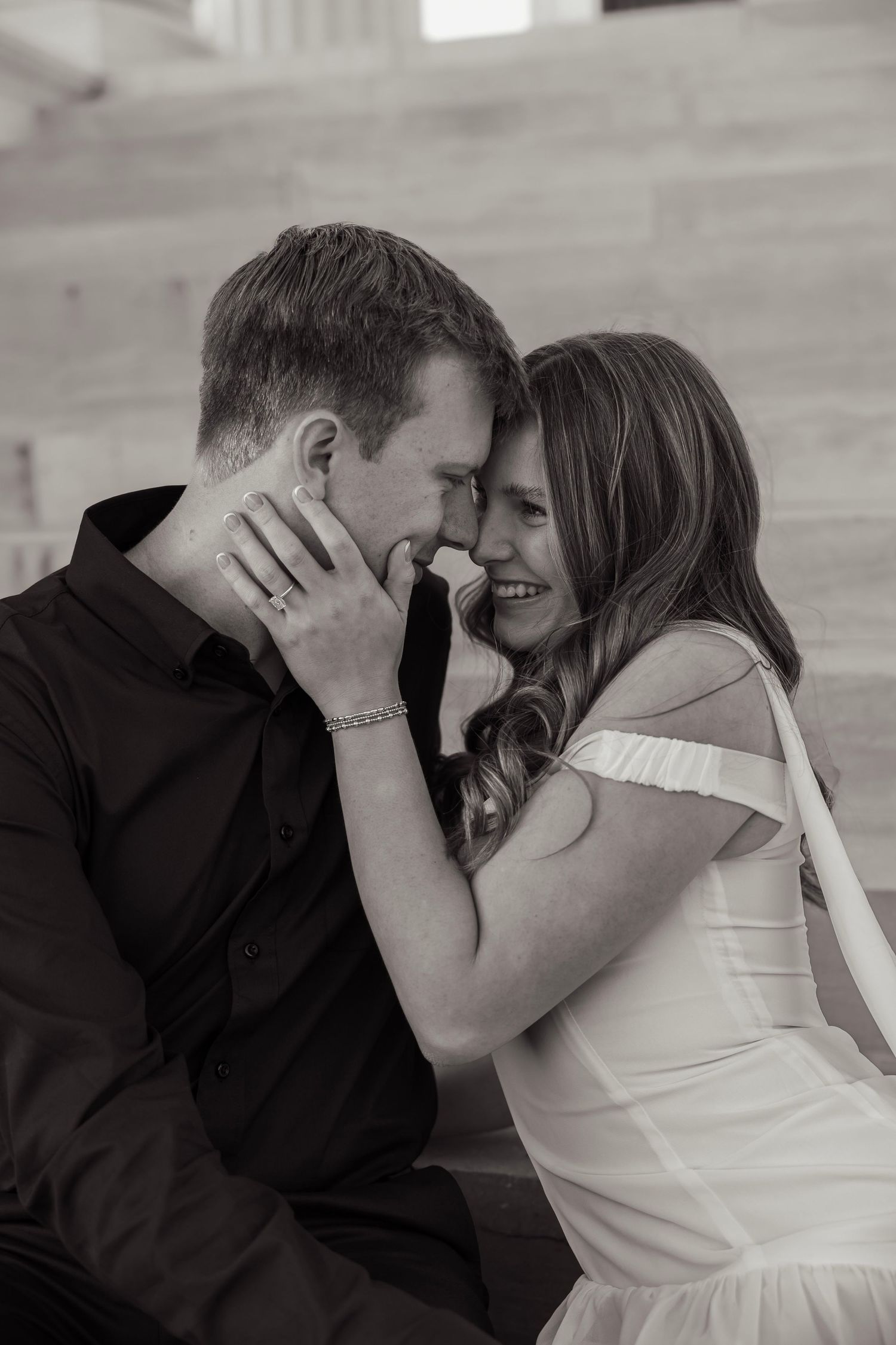 Black and white close-up of smiling couple touching foreheads tenderly on stone steps.