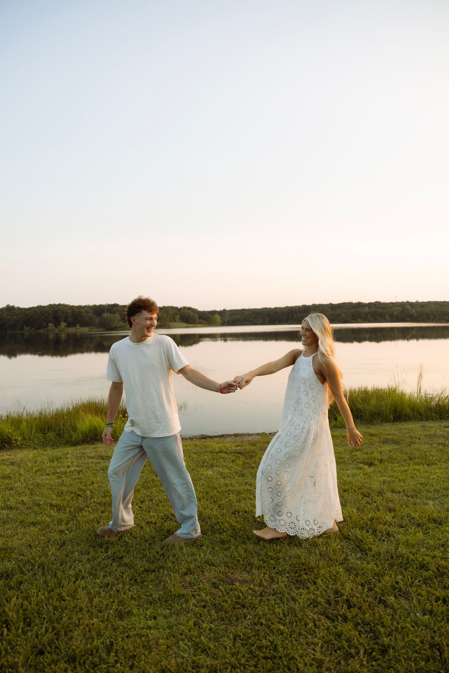 Couple holding hands and dancing barefoot on green grass by a serene lake at sunset.