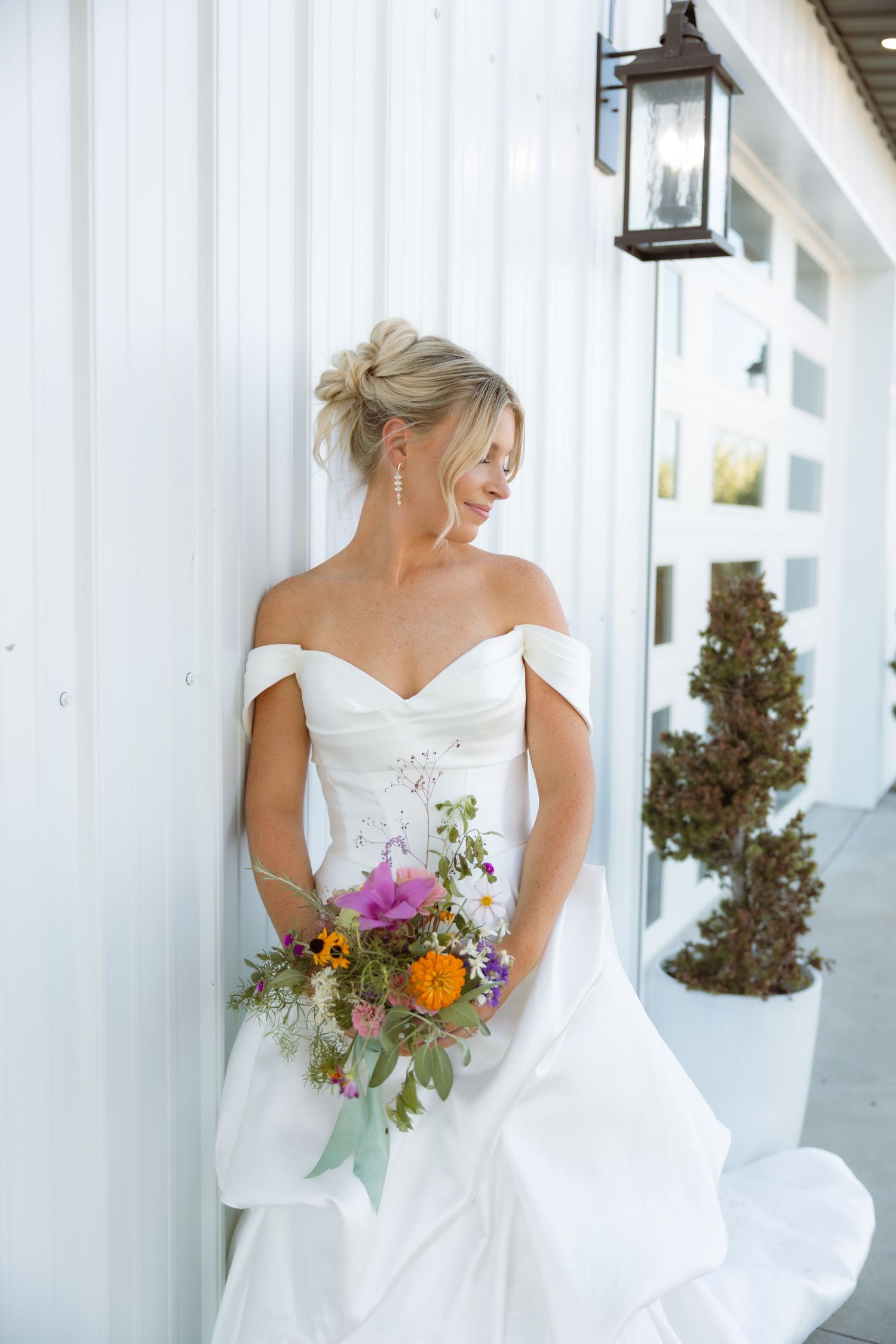 Bride in off-shoulder white gown holds colorful wildflower bouquet, leaning against white wall with vintage lantern.