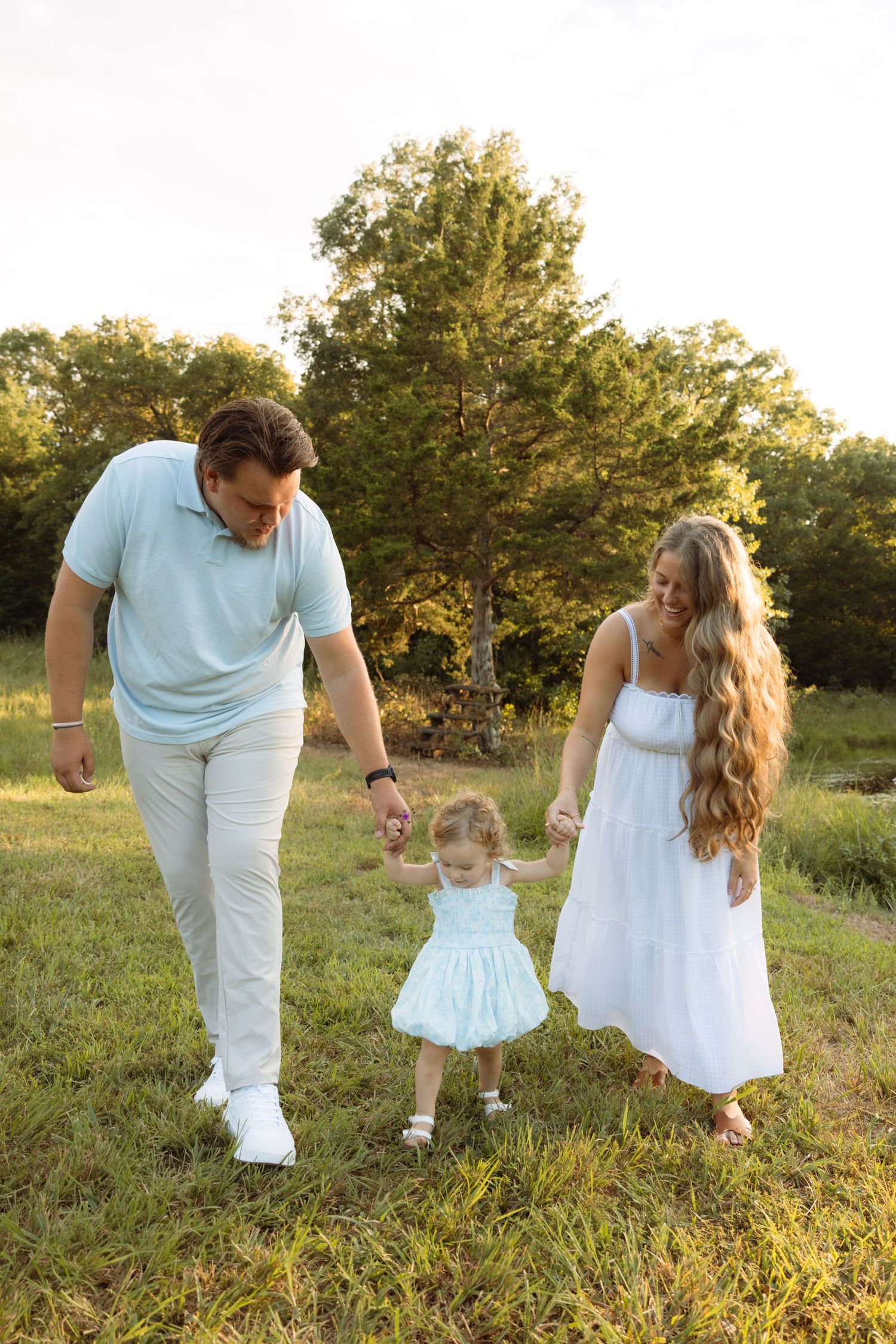 A young family walks hand-in-hand in a sunny green field, parents in white and blue outfits guiding their toddler daughter.