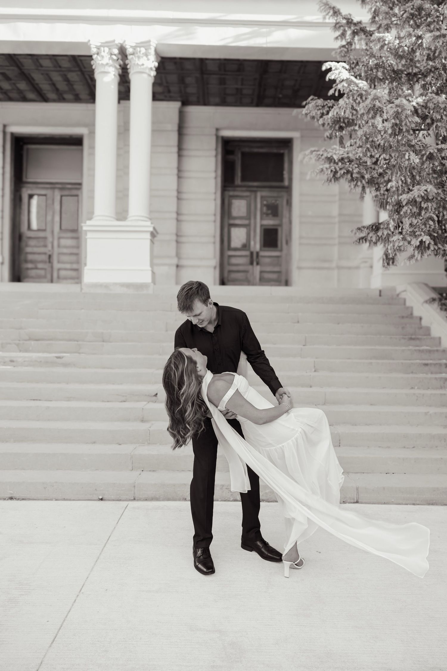 Black and white photo of groom dipping bride in front of a grand building with columns and steps.