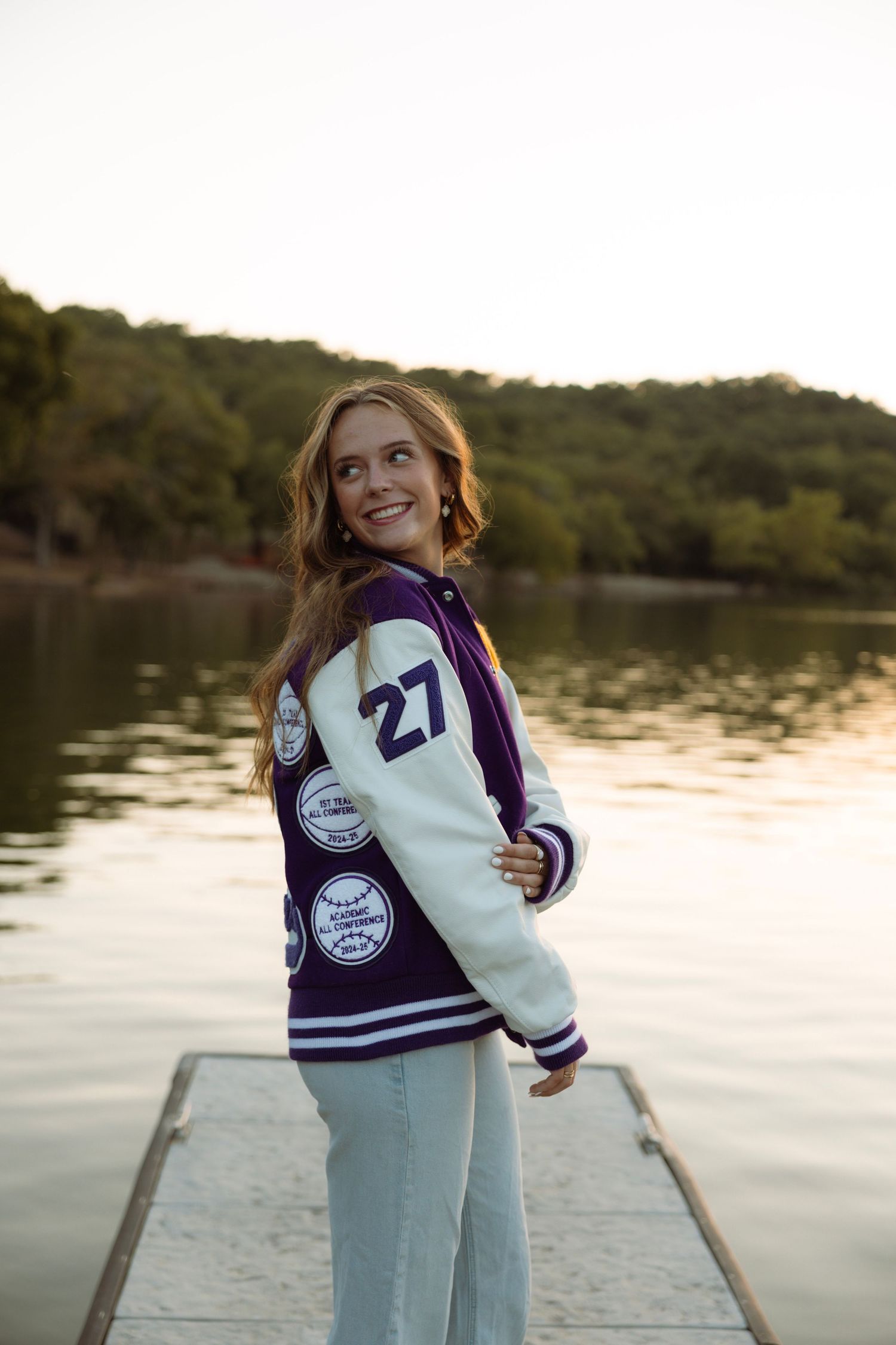 Young woman smiling in purple varsity jacket number 27 standing on dock by lake at sunset.
