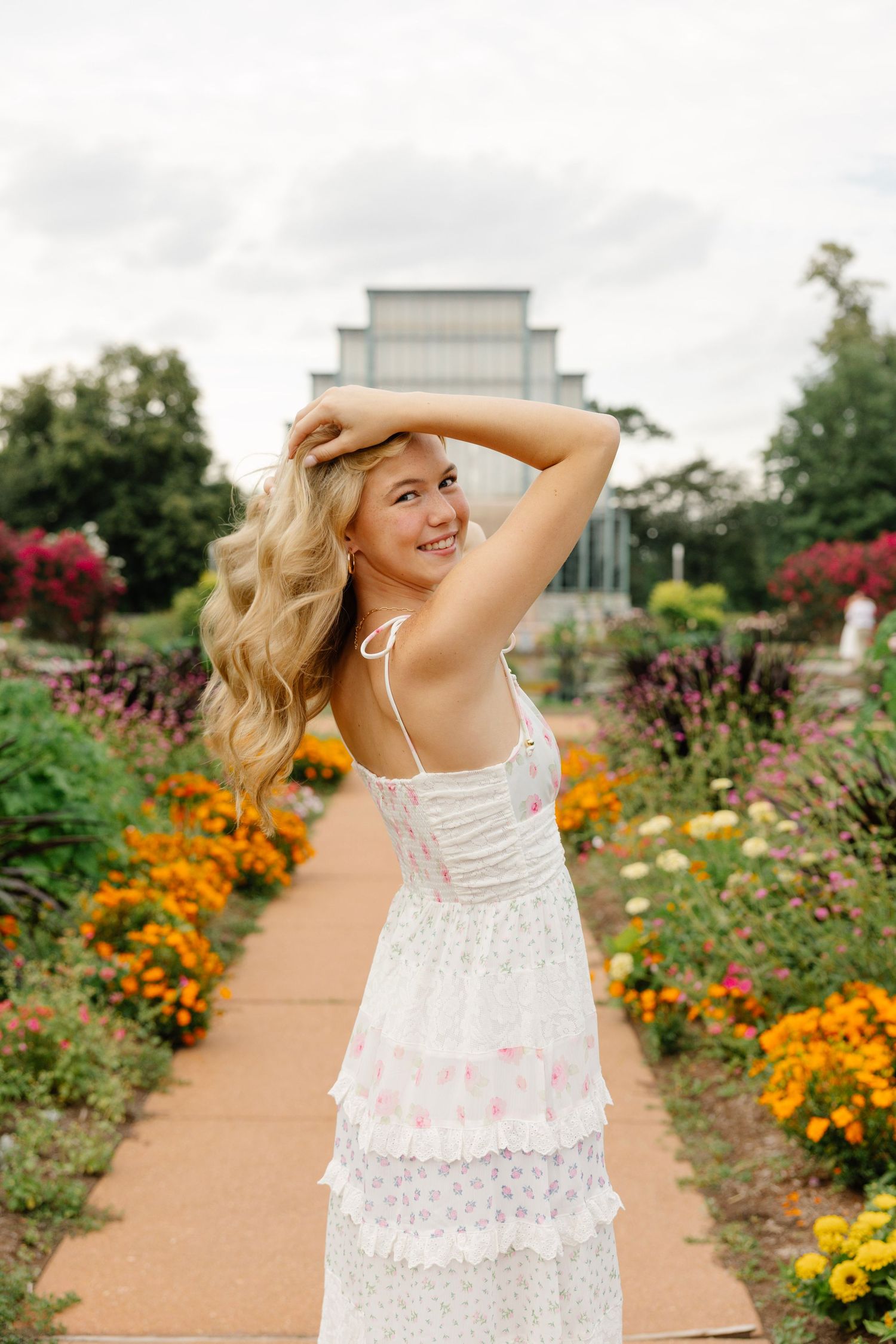 Young blonde woman in white floral dress poses in garden pathway surrounded by colorful flowers near a greenhouse.