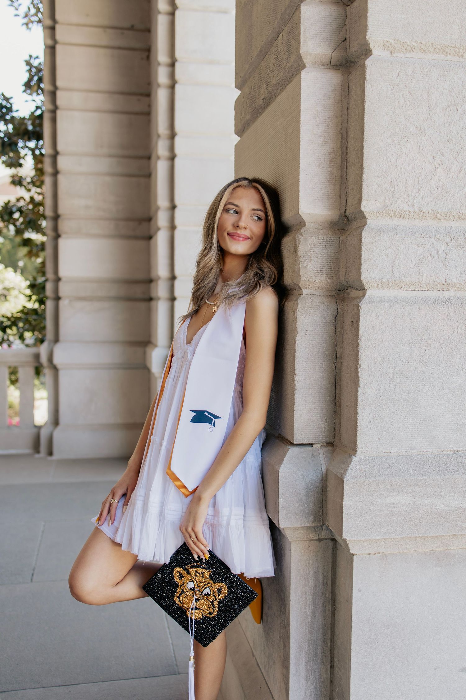 Female graduate in white dress leaning on stone column holding decorated tiger graduation cap.