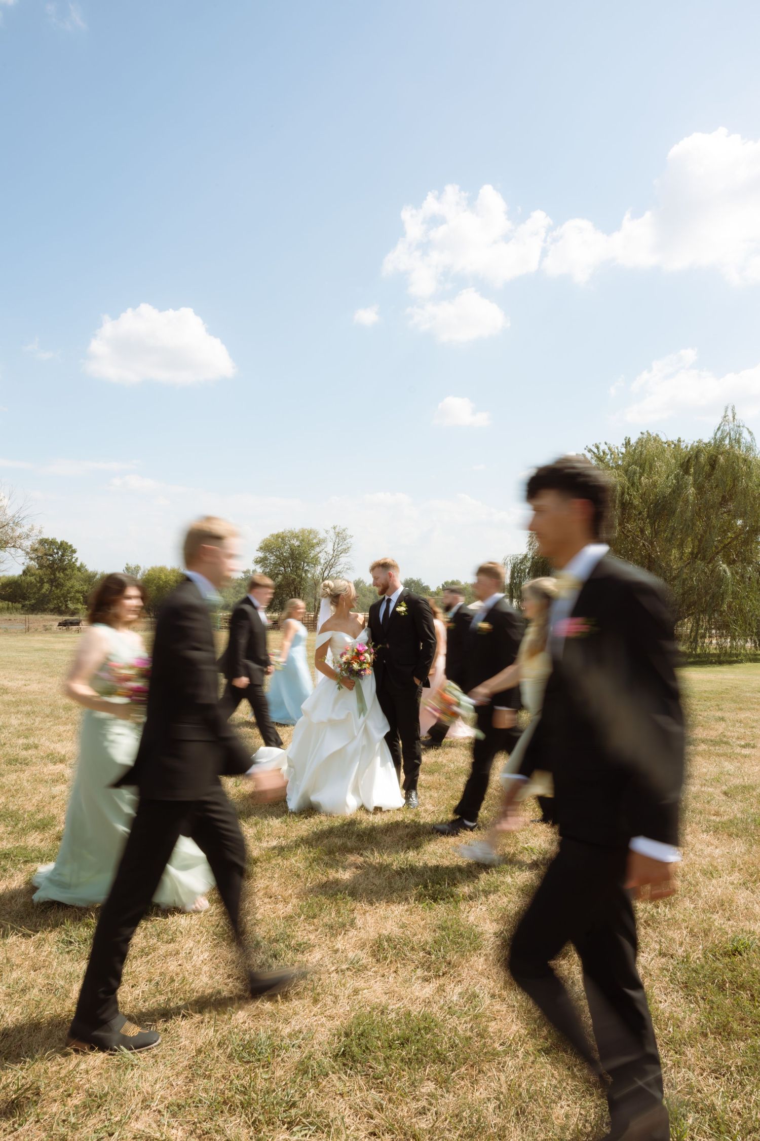 Bride and groom walk with wedding party on sunny green lawn, motion blur capturing joyful celebration outdoors.