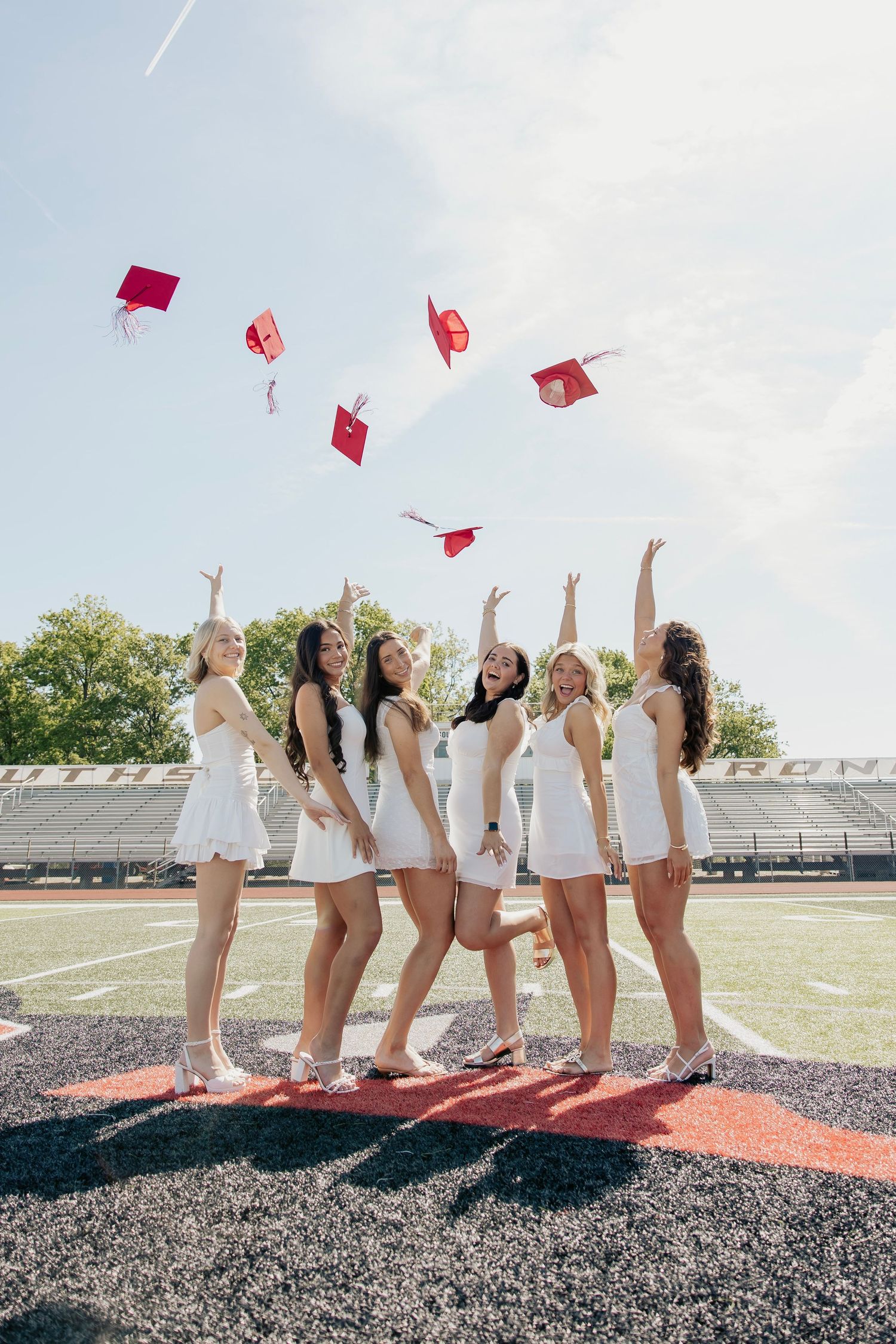 Six graduating girls in white dresses tossing red graduation caps in the air on a school football field.