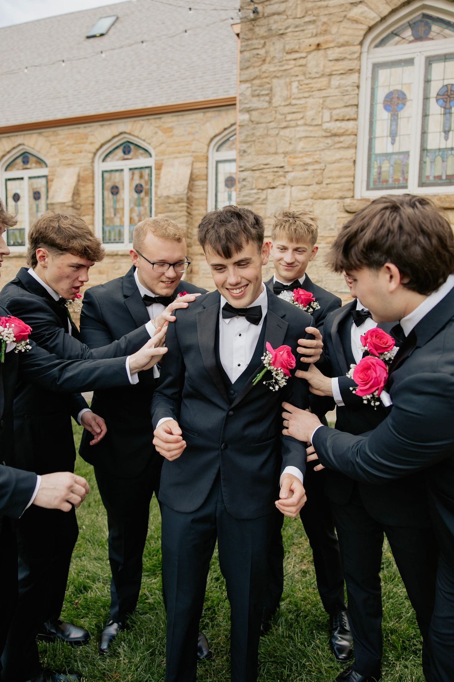 Groom in black tuxedo laughing with groomsmen wearing pink rose boutonnieres outside stone church.