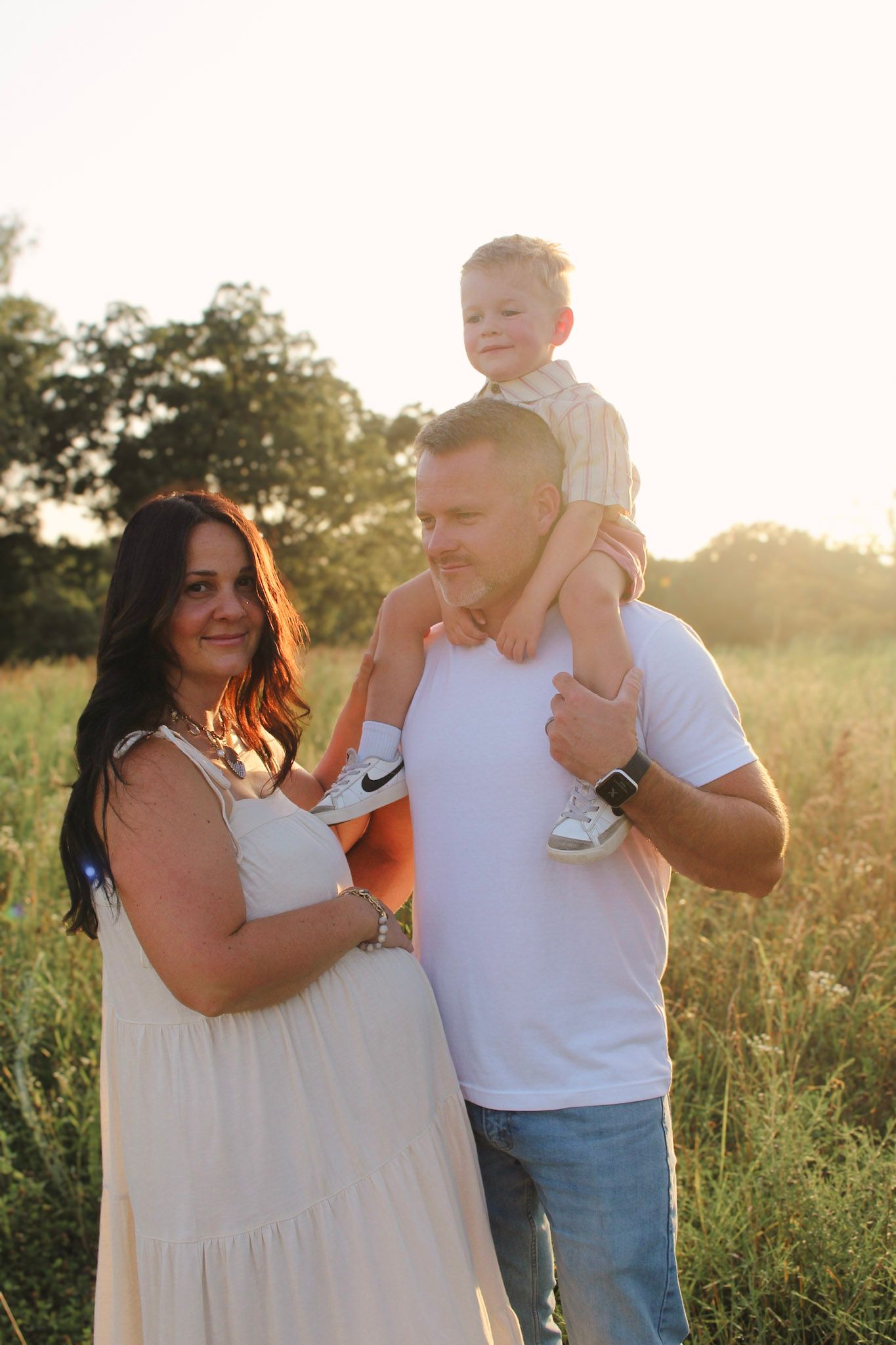 Smiling family portrait at sunset in a field, with young child sitting on fathers shoulders.