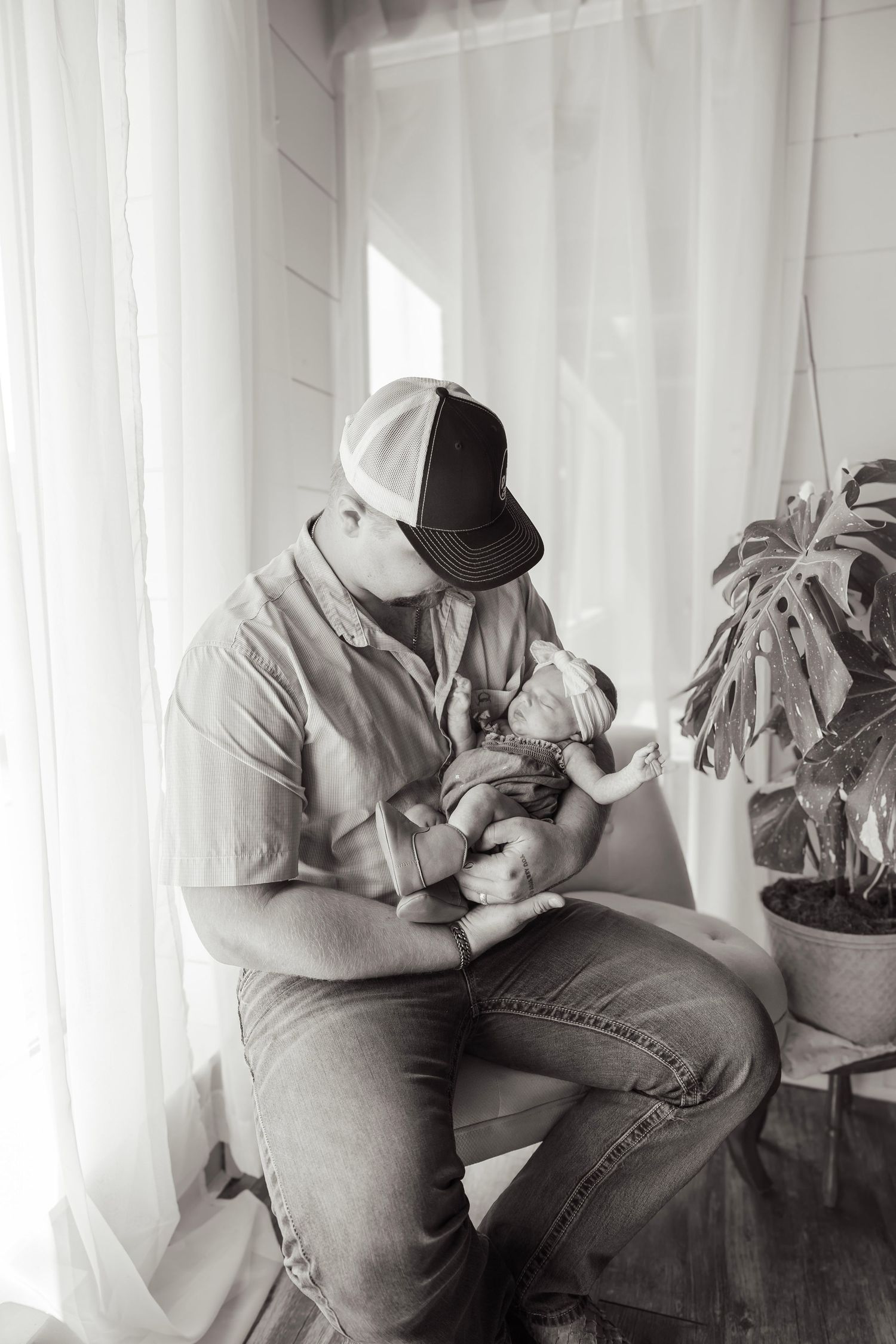 Black and white photo of a parent in a cap cradling a newborn baby near a sunlit window with sheer curtains.