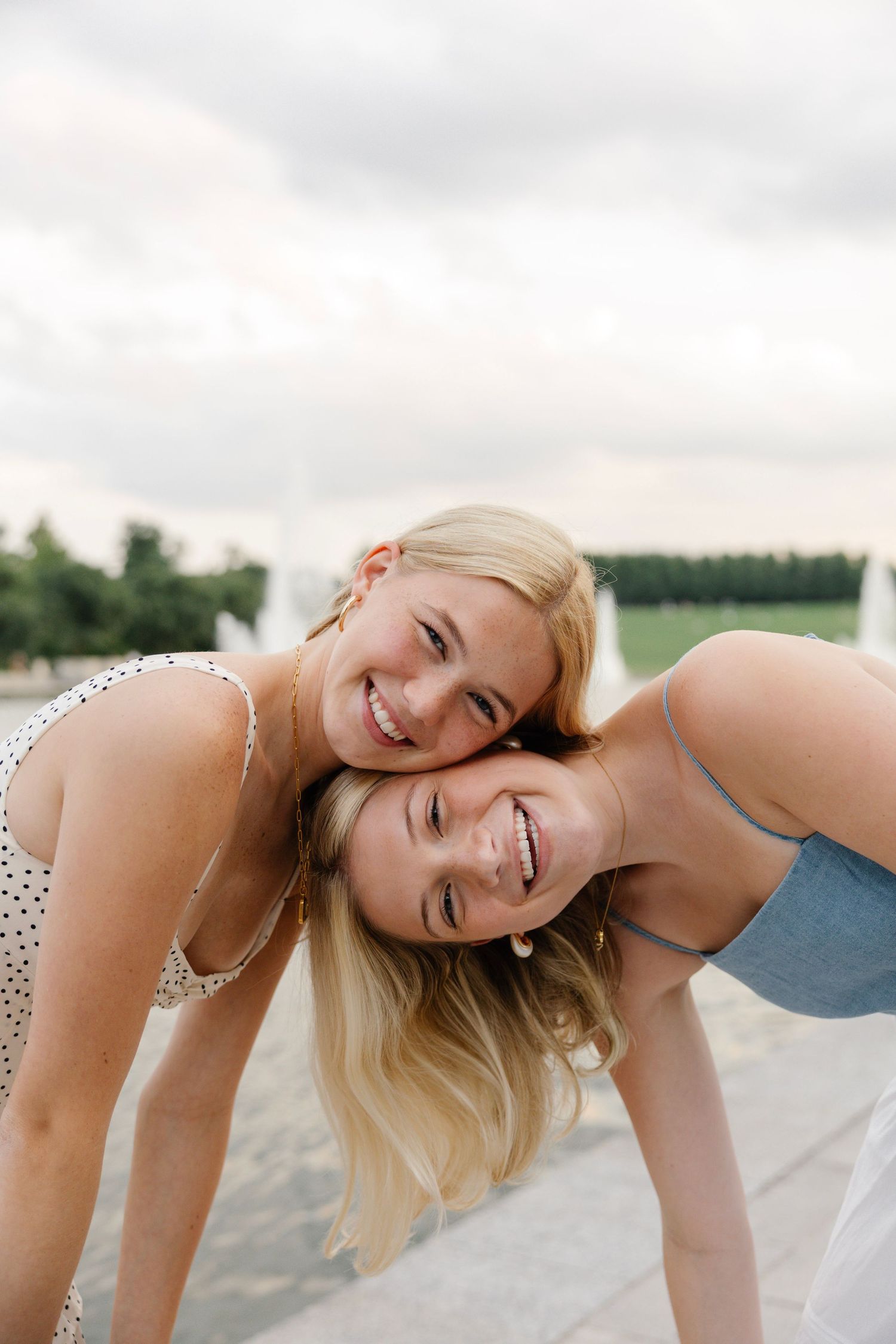 Two smiling blonde girls leaning heads together outdoors near a fountain on a cloudy day.