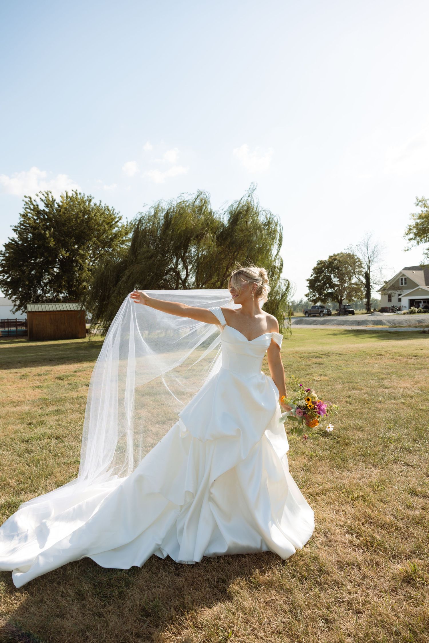 Bride in white off-shoulder gown holding flowing veil and colorful bouquet outdoors on sunny day.