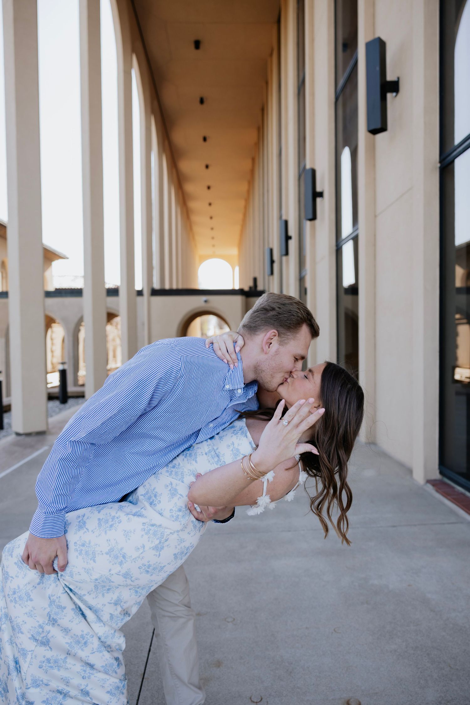 Couple sharing a passionate dip kiss in a sunlit columned outdoor corridor.