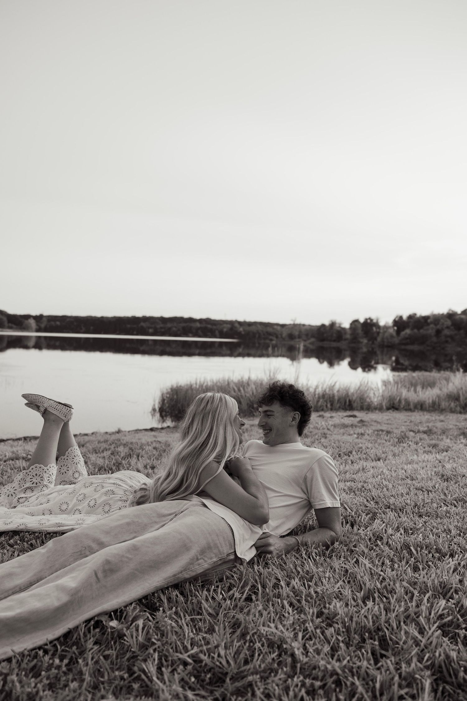 Black and white photo of a couple lying on grass by a lake, smiling at each other in a romantic outdoor setting.