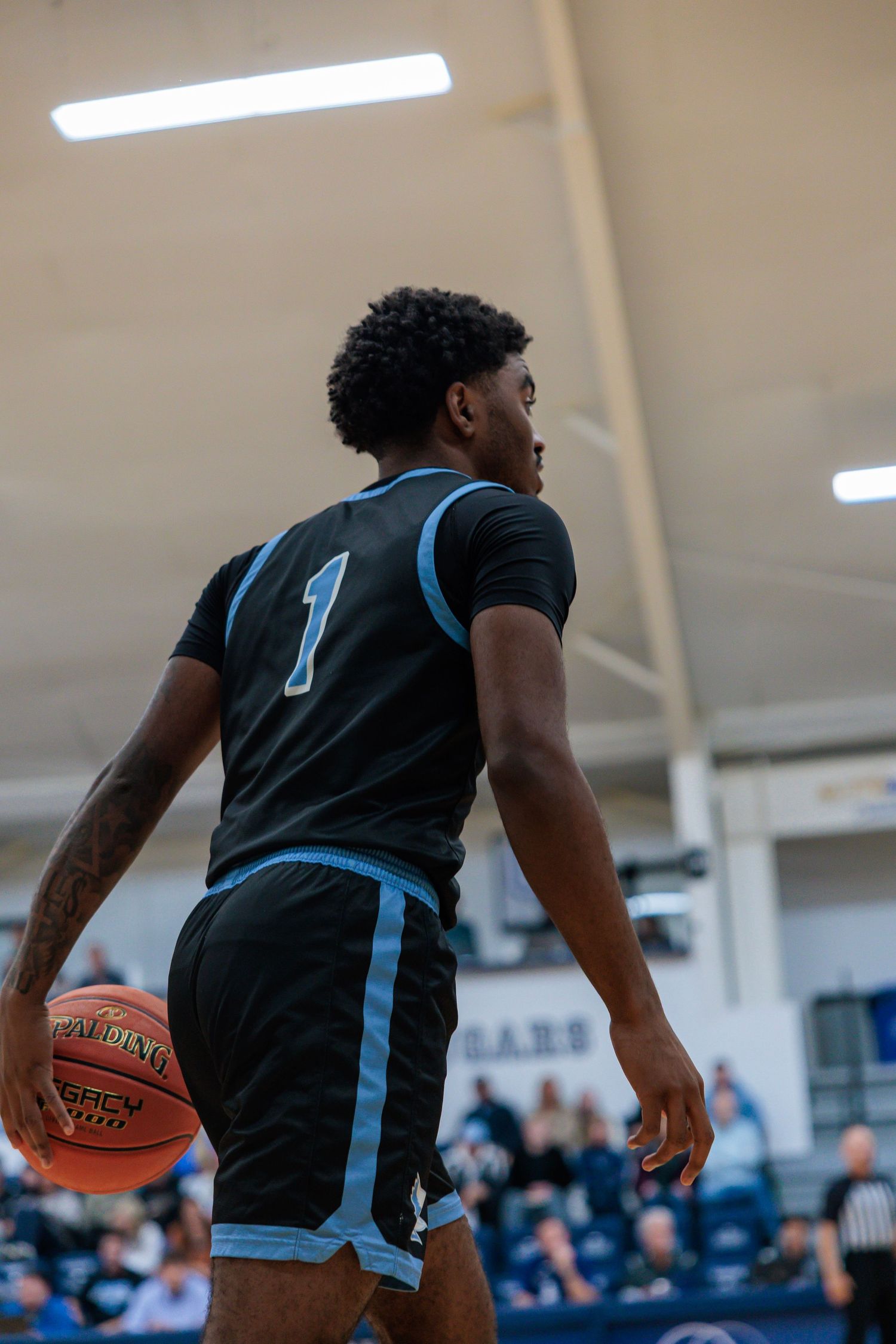 Basketball player wearing number 1 dark jersey dribbling ball viewed from behind during an indoor college game.