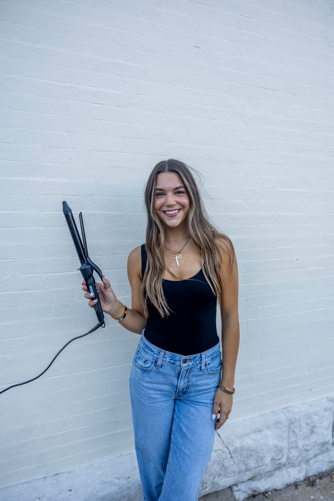 A smiling young woman in a black bodysuit and jeans holds a curling iron against a white brick wall.