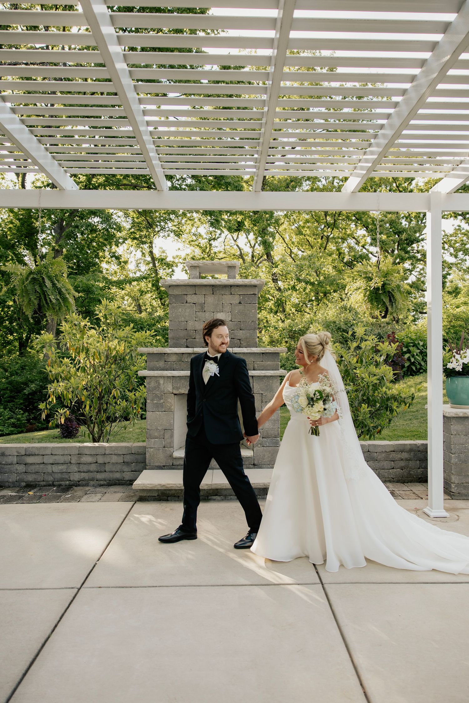 Bride and groom hold hands under a white pergola surrounded by lush greenery on their wedding day.