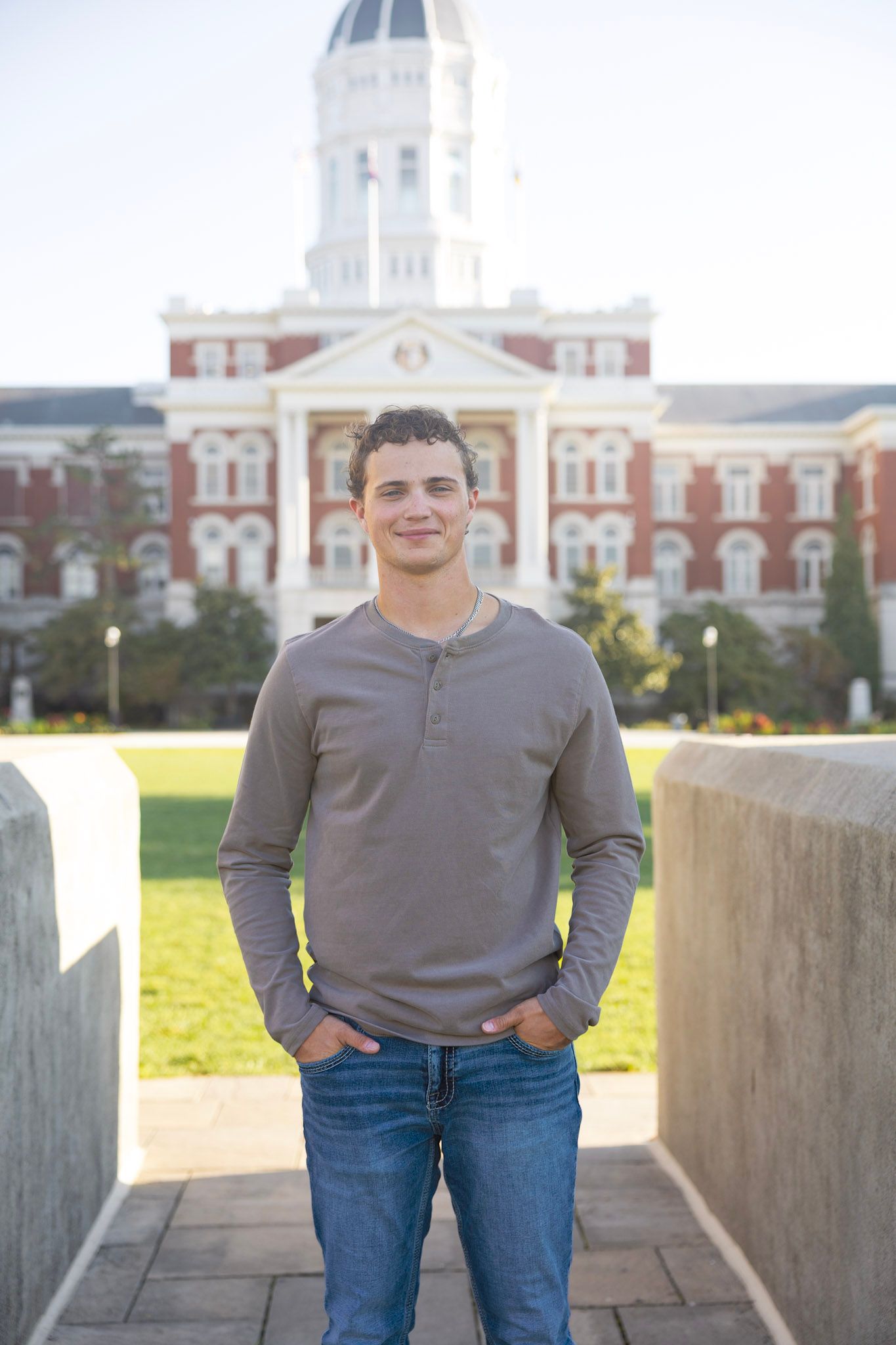 Smiling young man in a gray henley standing in front of a historic red brick university building.