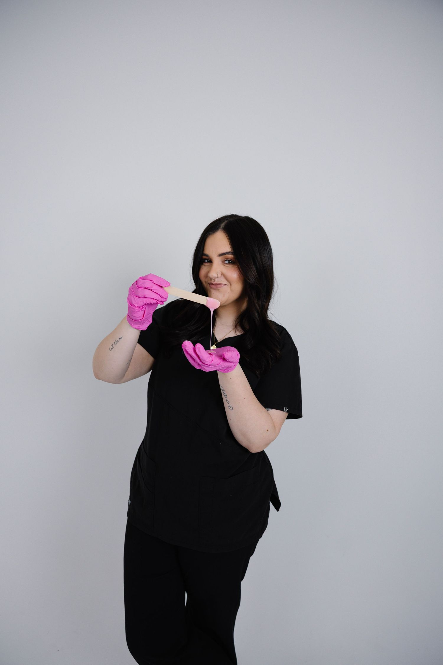 Female esthetician in black scrubs and pink gloves holding a wooden wax applicator stick.