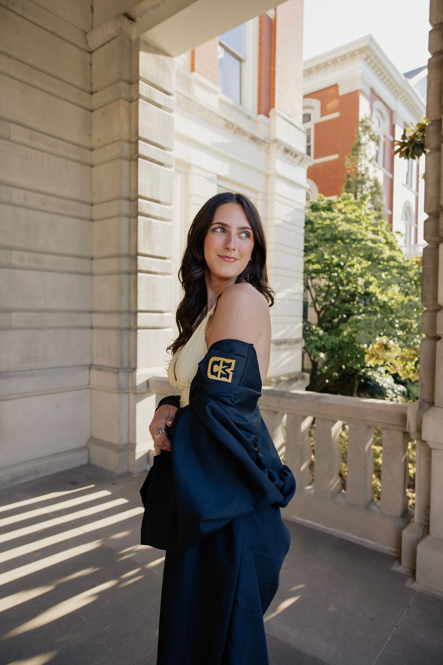 Brunette female graduate in off-shoulder top holding navy graduation gown on sunlit balcony.