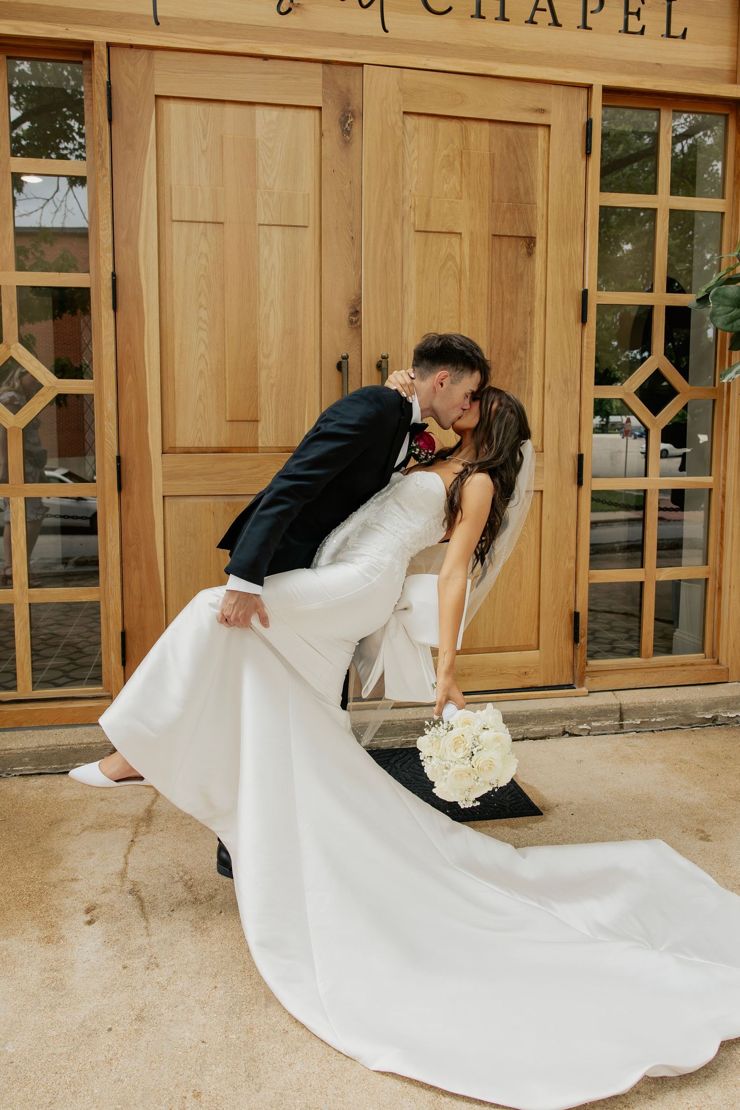 Groom dipping bride for a kiss outside wooden chapel entrance doors.