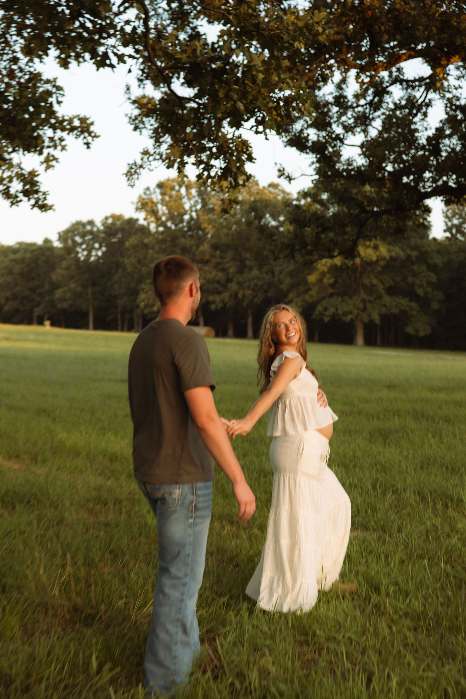 Pregnant couple holding hands in a sunlit green field, woman in white dress glancing back, trees in background.