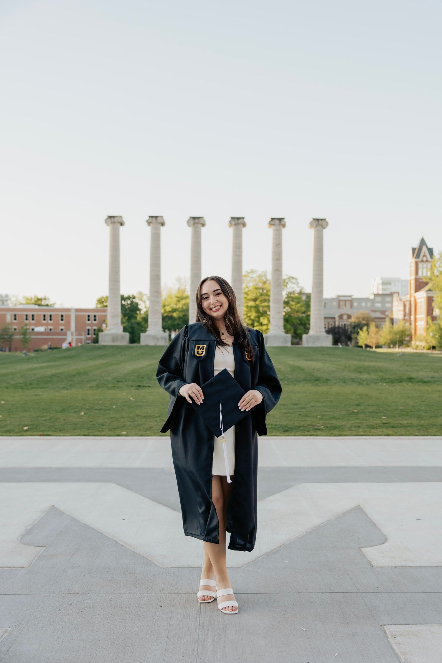Smiling graduate in black cap and gown poses before the iconic columns at University of Missouri campus.