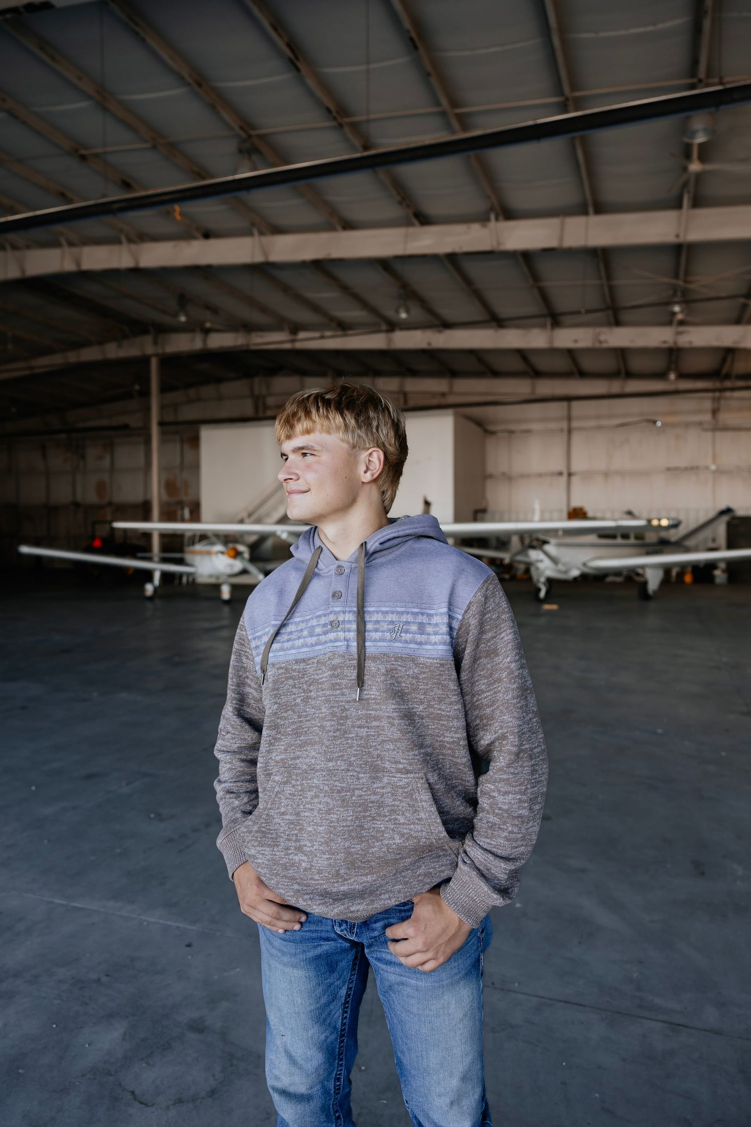 Smiling teen boy in gray hoodie standing in airplane hangar.