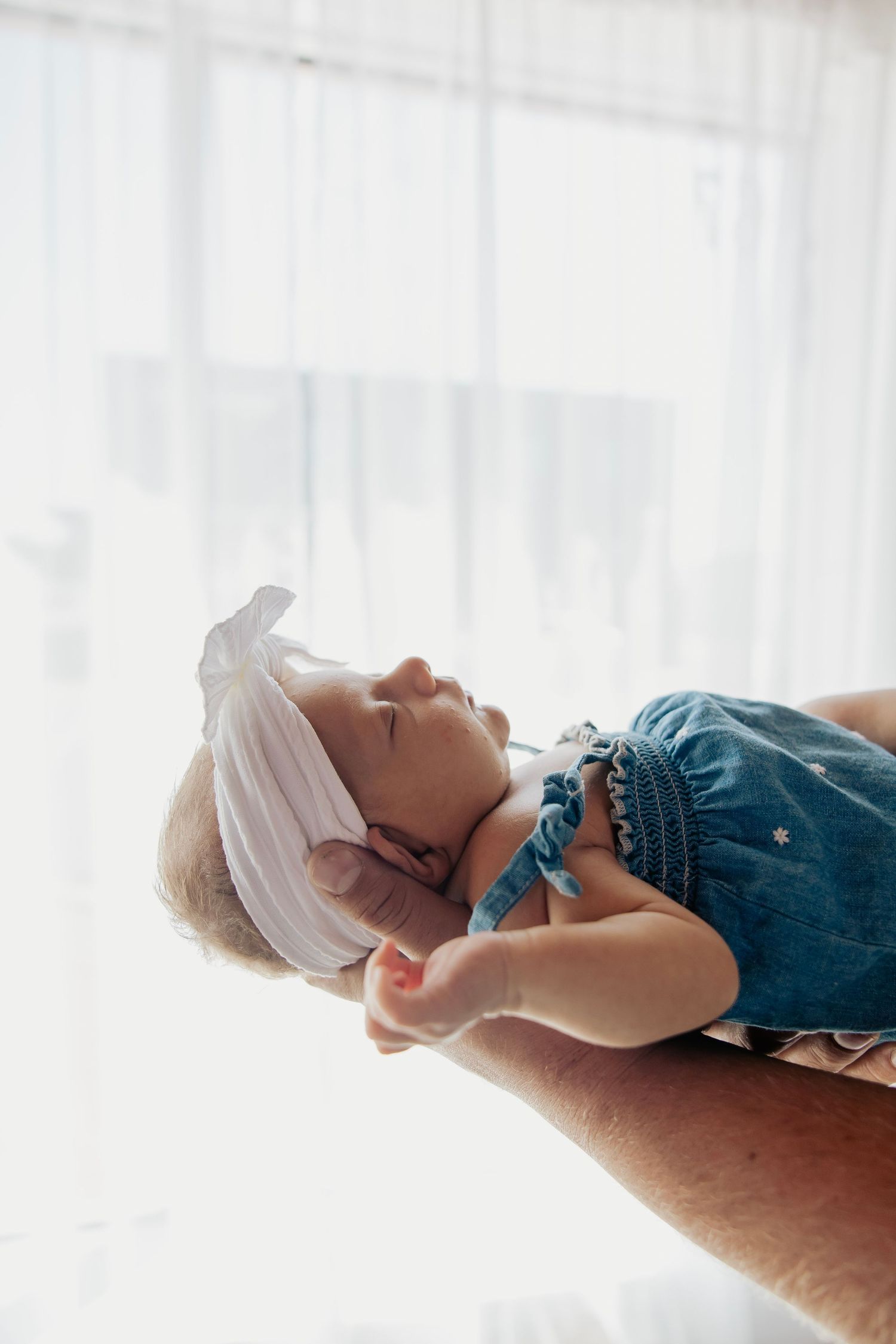 A sleeping baby in a denim romper and white bow headband being cradled in adult hands near a bright window.