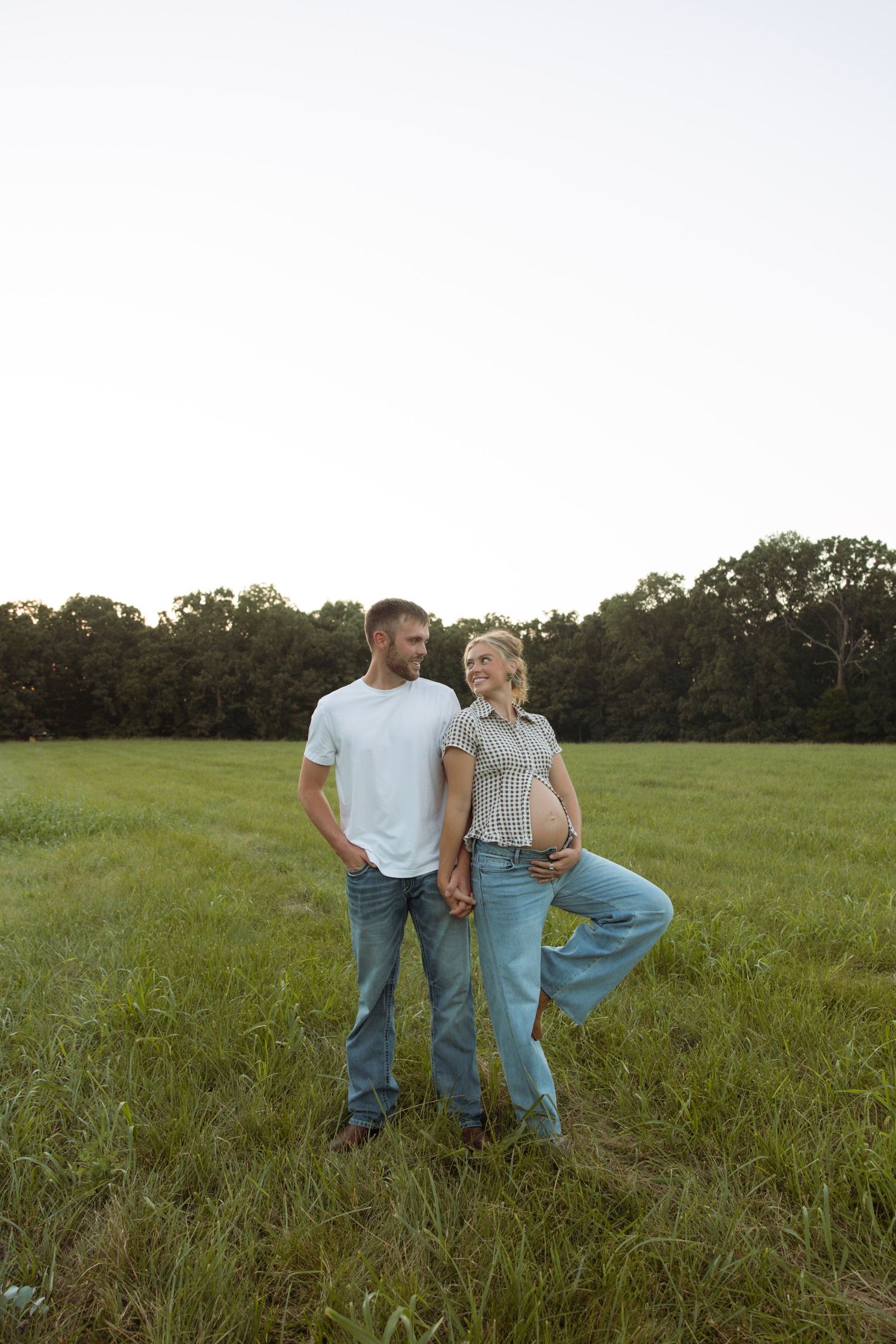 A couple stands in a green field at sunset, the woman visibly pregnant, both smiling and holding hands.