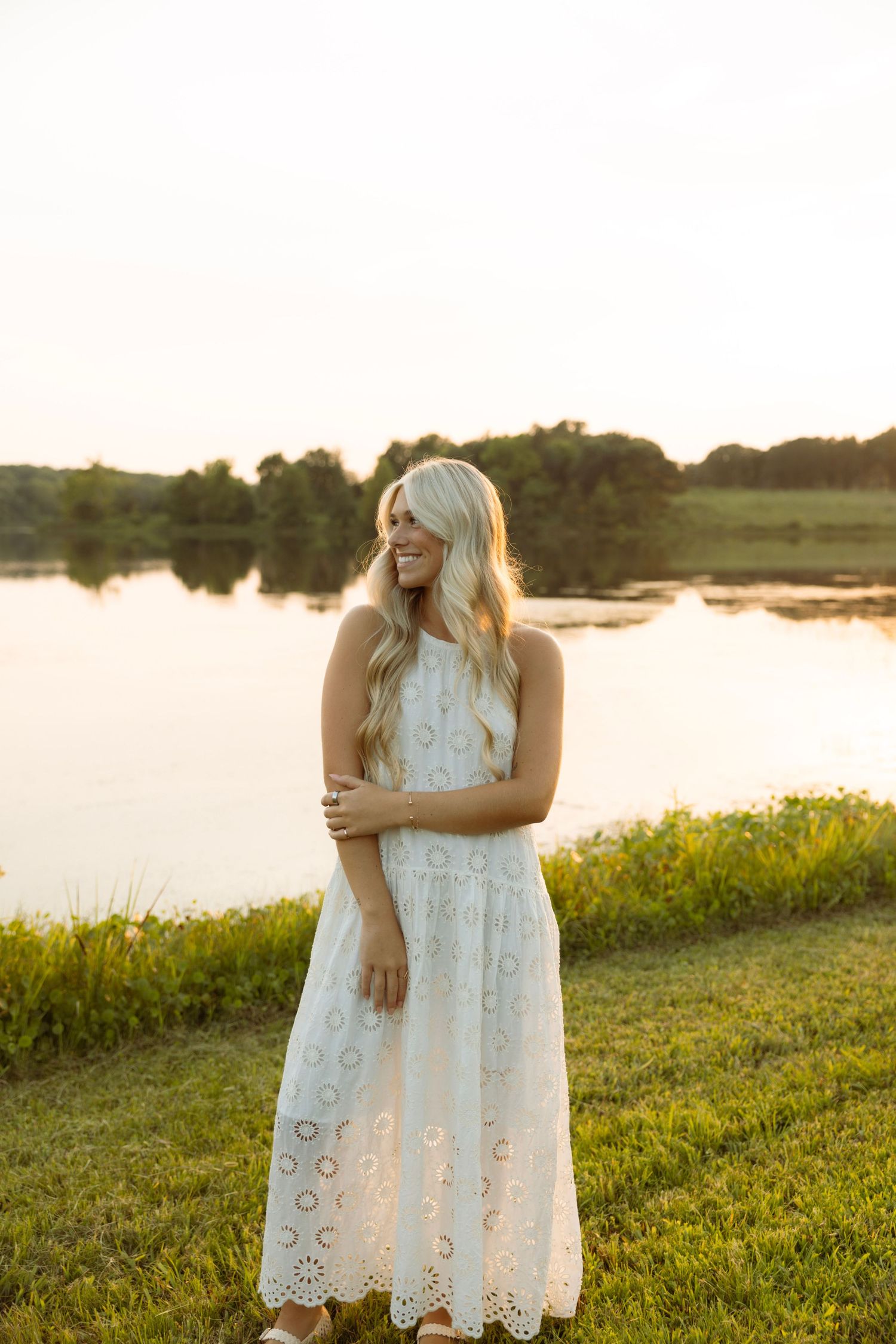 Smiling blonde woman in white eyelet maxi dress standing by a serene lake at golden hour sunset.