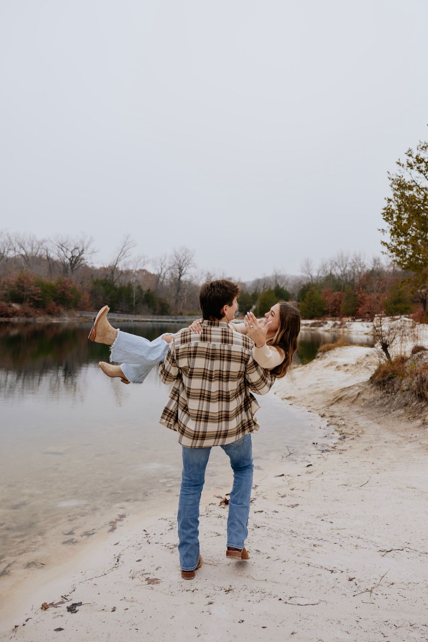 A couple shares a romantic moment as a man carries a woman in his arms along a sandy riverbank surrounded by autumn trees.