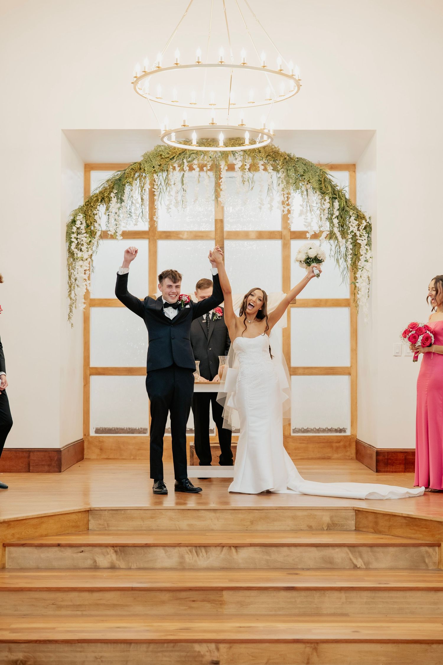 Joyful newlyweds raise their arms in celebration at wedding altar adorned with greenery chandelier in elegant white venue.