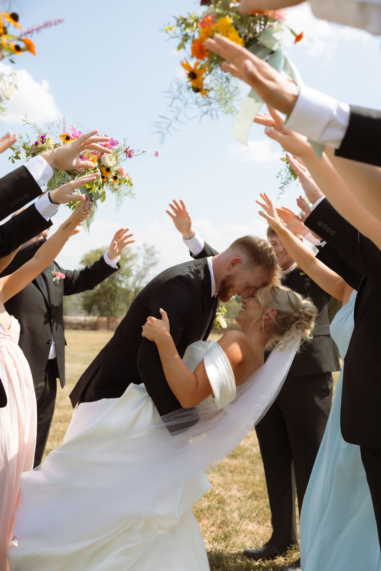 Bride and groom kissing under raised hands and floral bouquets of wedding party outdoors on sunny day.