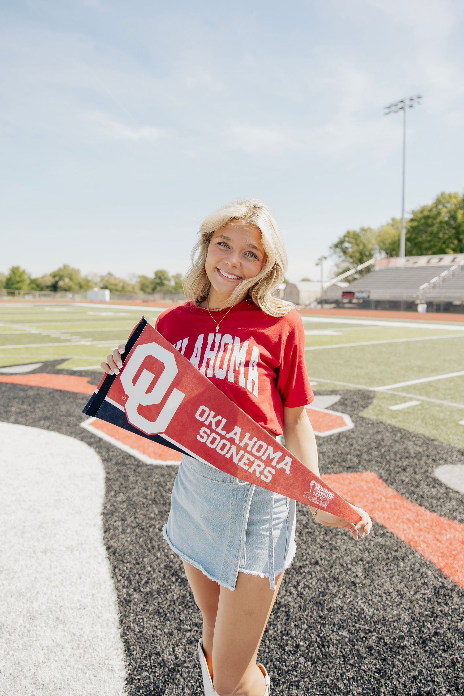 Smiling blonde girl holding Oklahoma Sooners pennants on a football field wearing a red OU t-shirt.