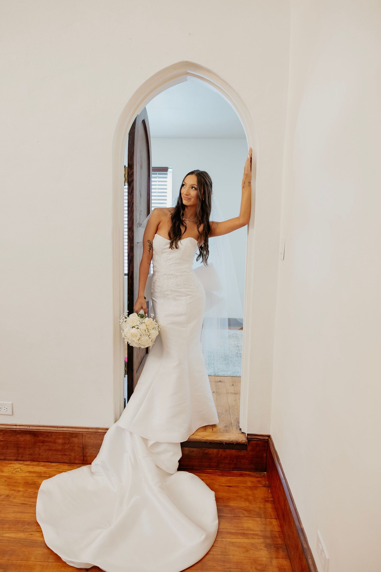 Elegant bride in mermaid wedding dress posing in arched doorway holding white bouquet.